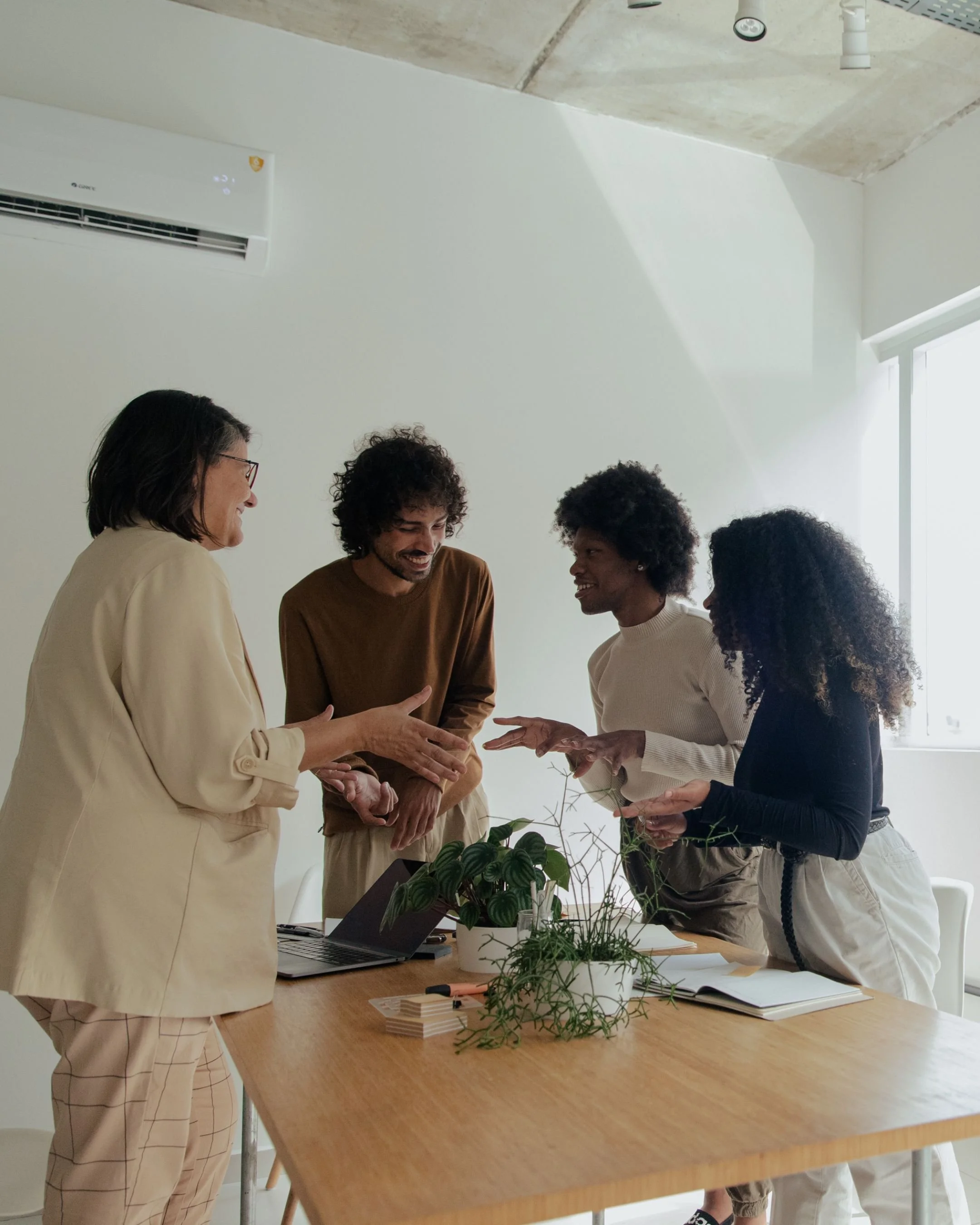 Four diverse colleagues having a lively discussion around a wooden table in a bright office, with a laptop and plants on the table.