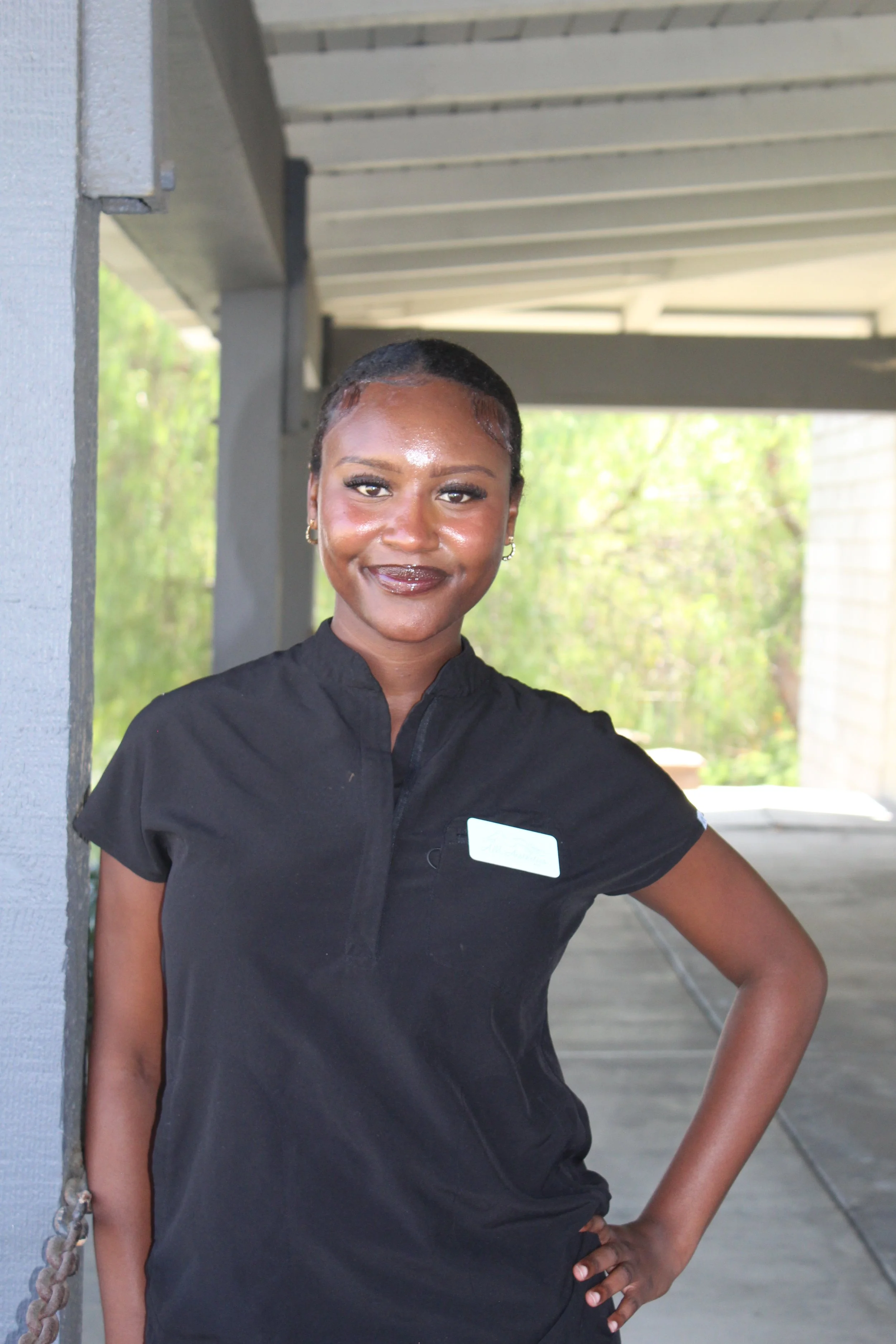 Portrait of a woman with dark skin, short black hair, wearing a black uniform with a white name tag, standing outdoors under a covered area with green trees in the background.