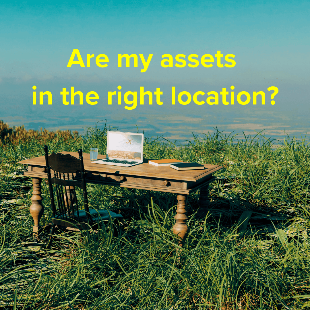 Title: "Are my assets in the right location?" A brown wooden desk with a black wooden chair sit on a hill amid tall grasses overlooking a valley. On the table are a laptop, a couple books, and a glass of water.