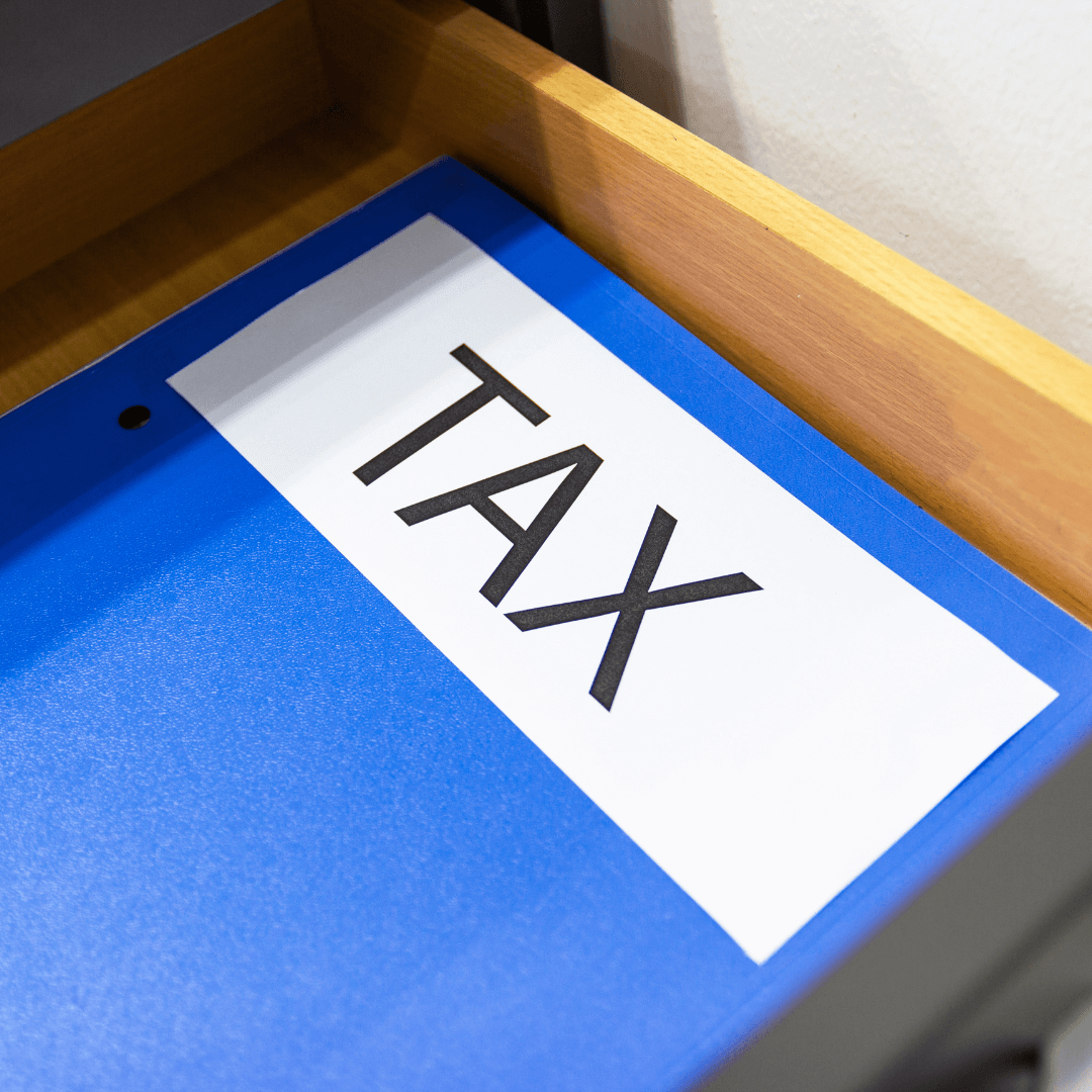 Photo of a blue folder with TAX written on it. The folder is within a wooden desk drawer which is next to a white wall.