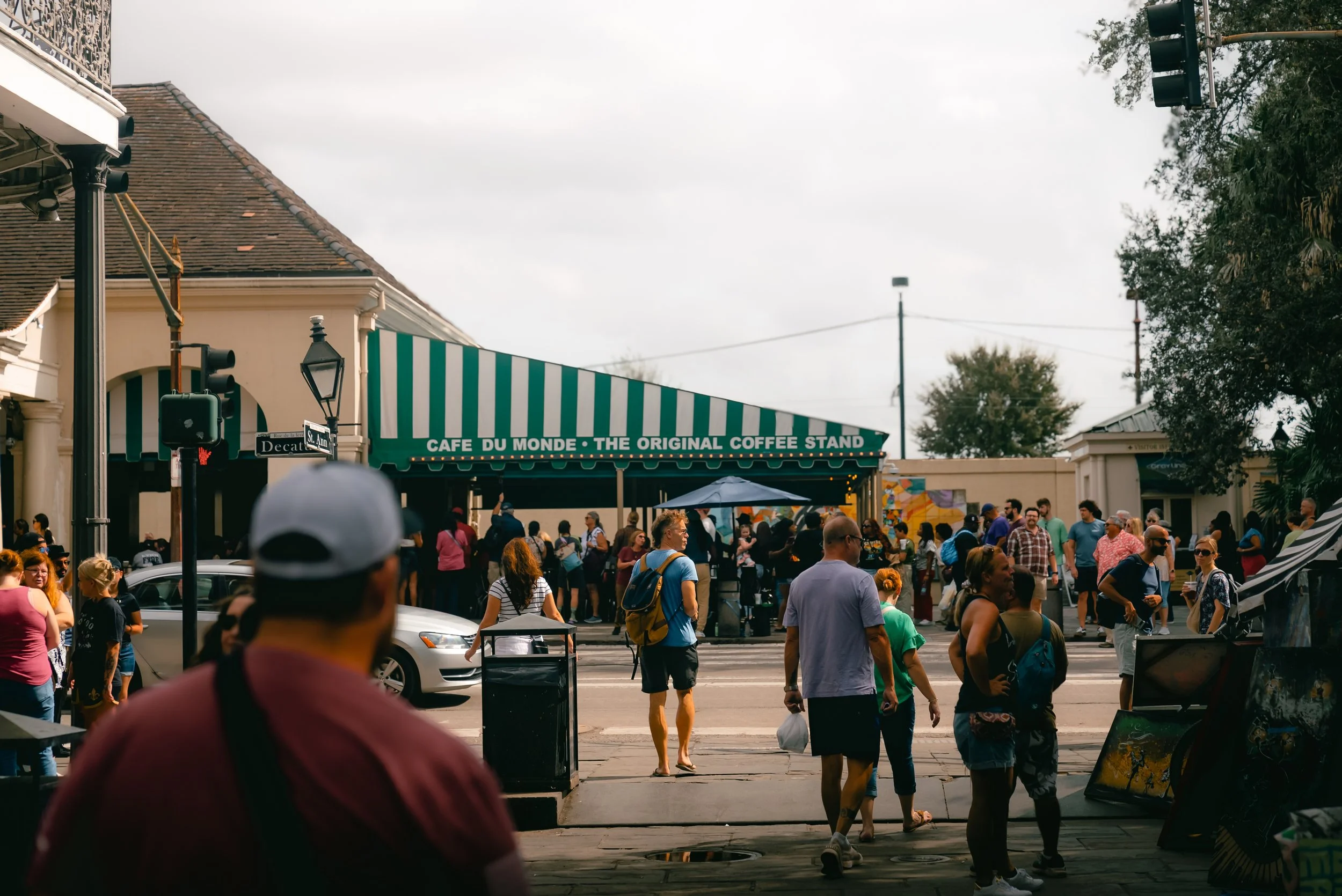 A busy street scene with people walking and standing. A green and white striped awning with the sign 'Cafe du Monde - The Original Coffee Stand' is visible across the street. There are buildings on both sides and some trees in the background.