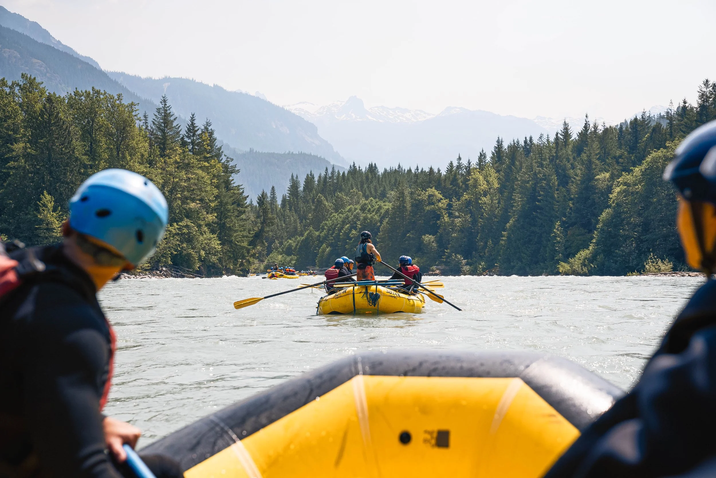 People rafting on a river surrounded by dense green forest and mountains in the background.