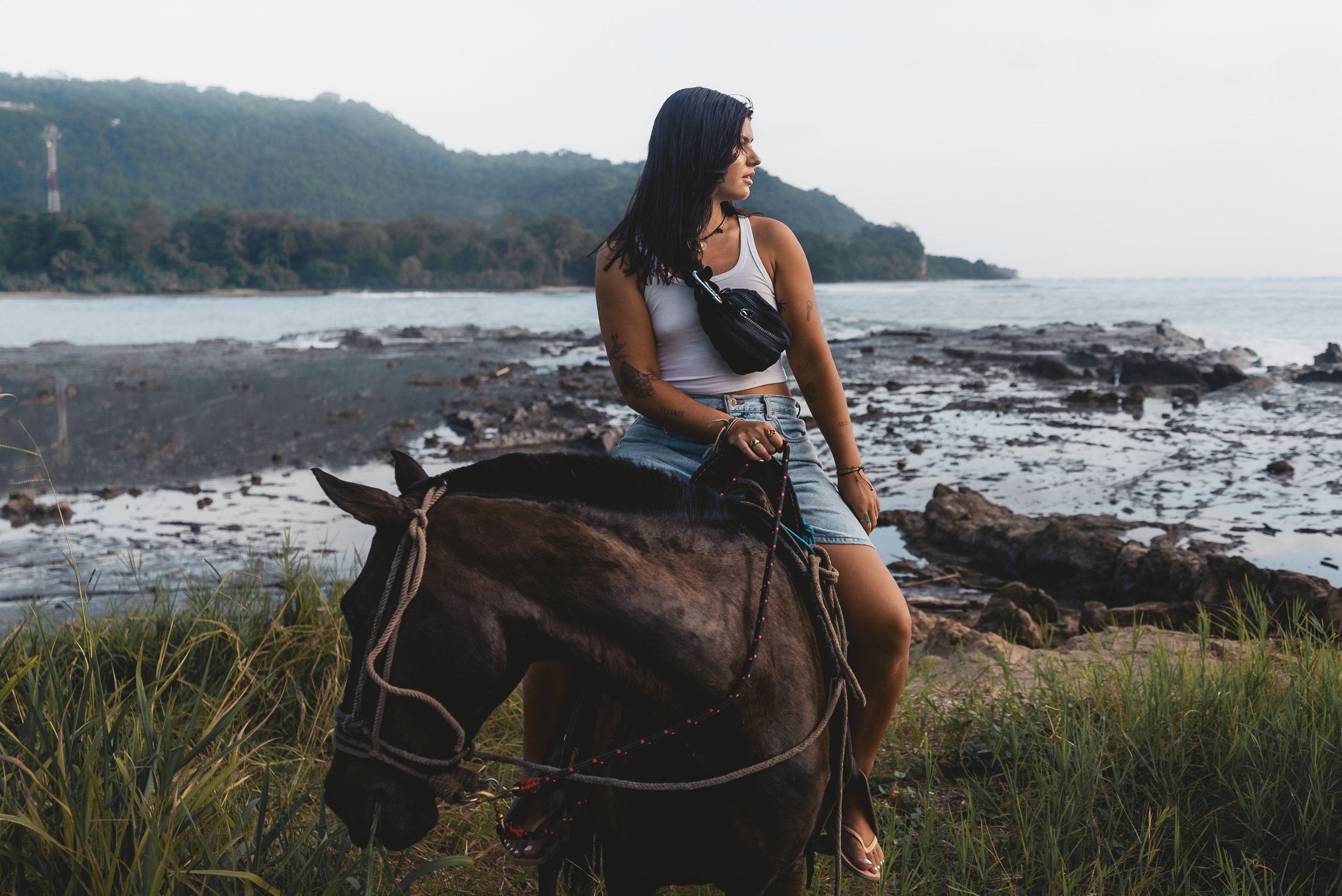 A woman with black hair wearing a grey tank top and denim shorts rides a horse along a rocky beach with water and green hills in the background.