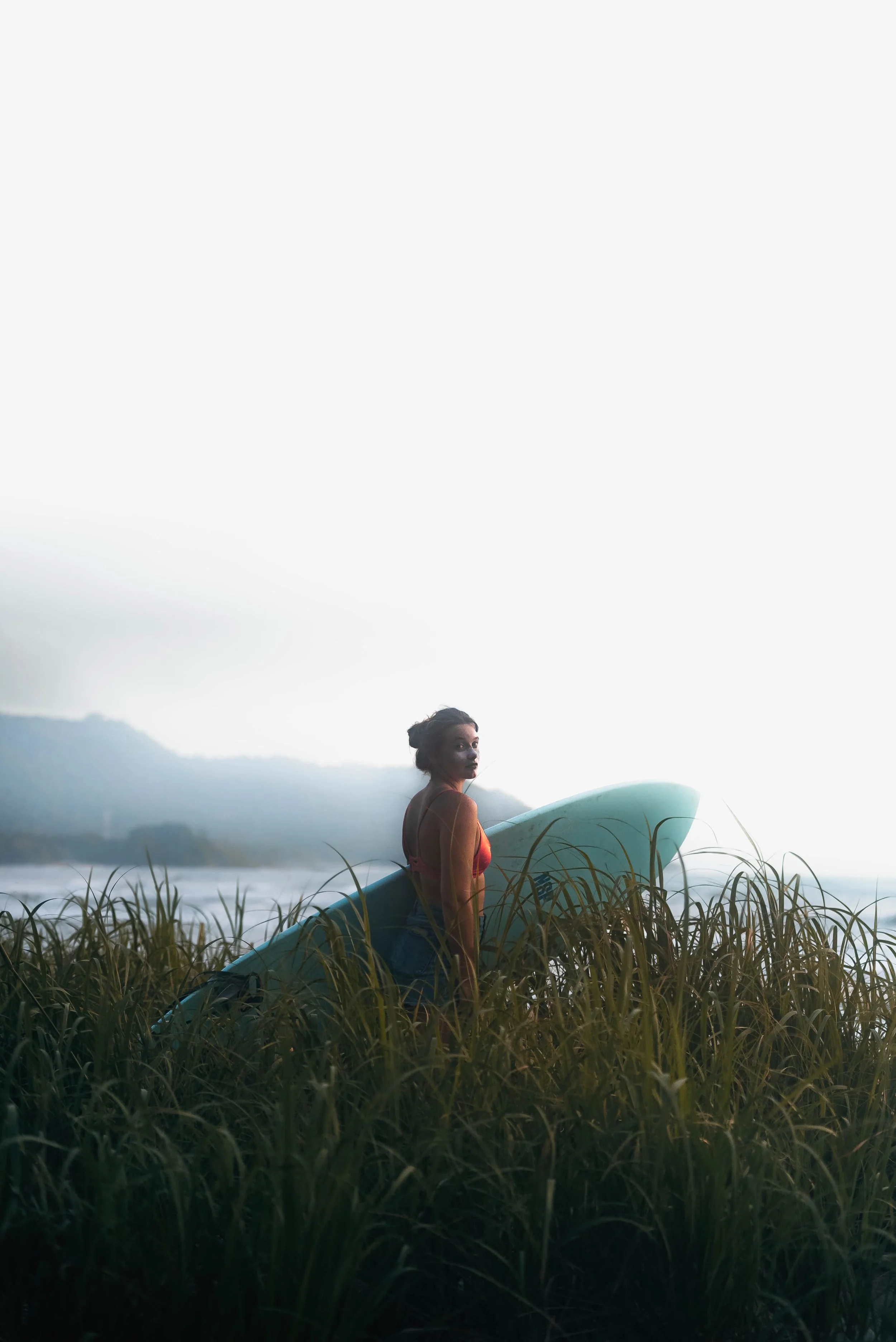 A woman with a surfboard standing in tall grass near a body of water during dusk or dawn, with mountains in the background.
