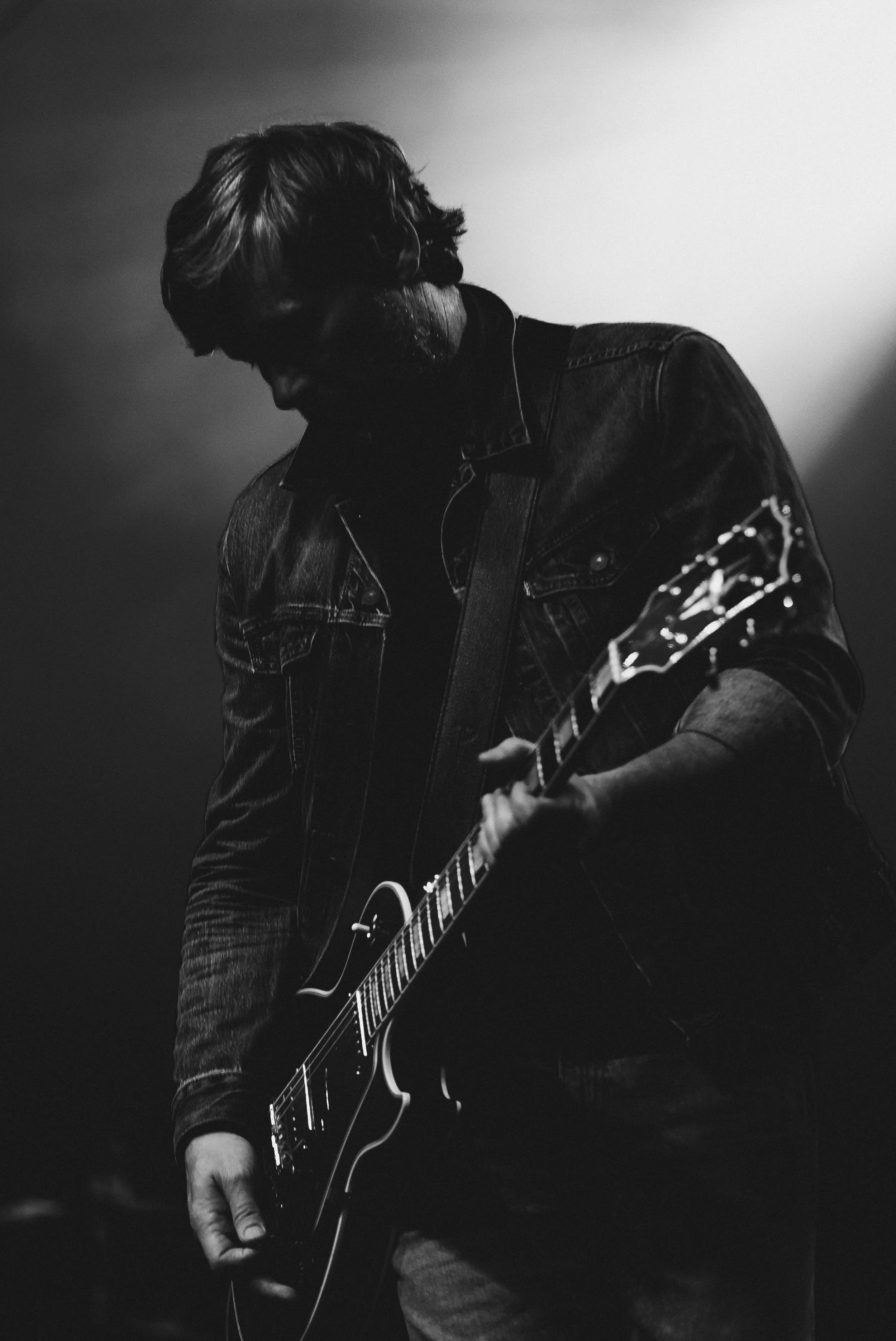 A man playing an electric guitar on stage, wearing a denim jacket and jeans, with his head bowed down in concentration.