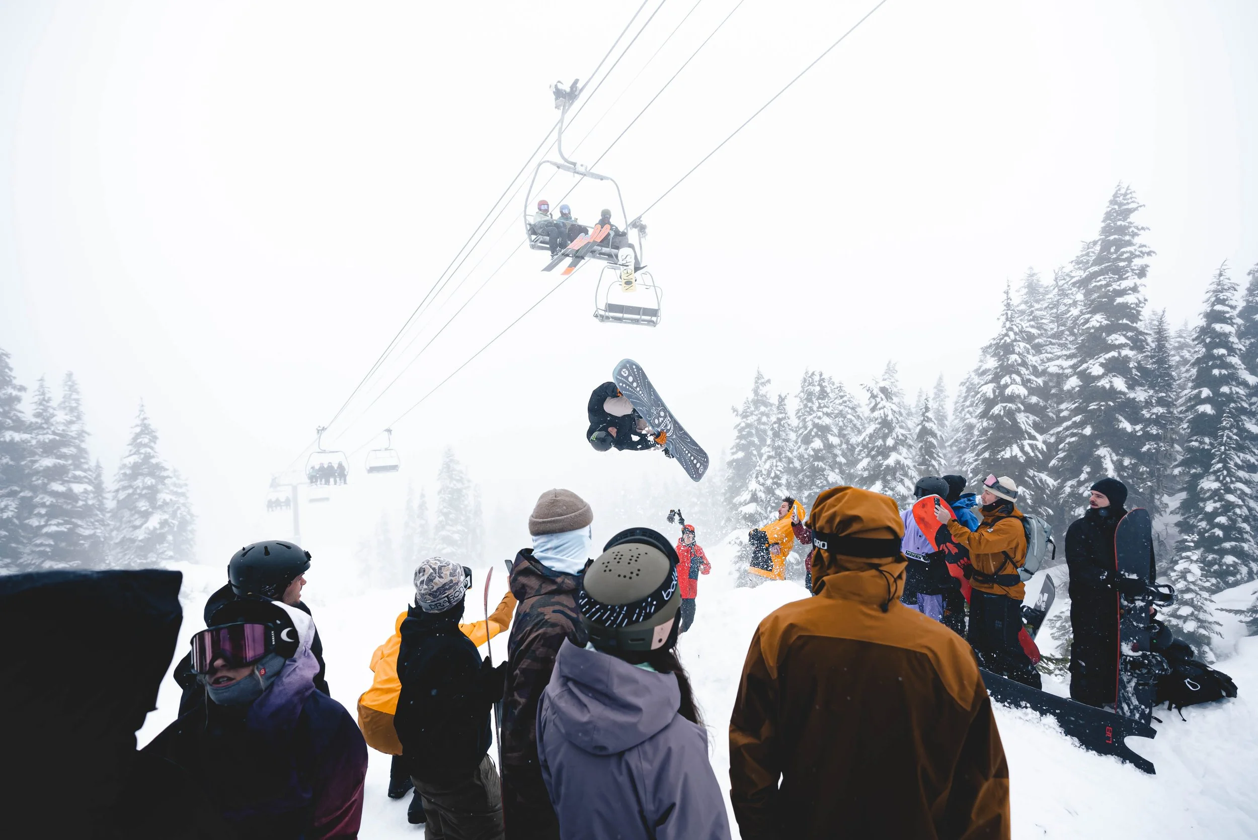 Group of people in winter clothing gathered at a snowy ski resort, with some skiing and snowboarding, and a ski lift in the background amid snow-covered trees.