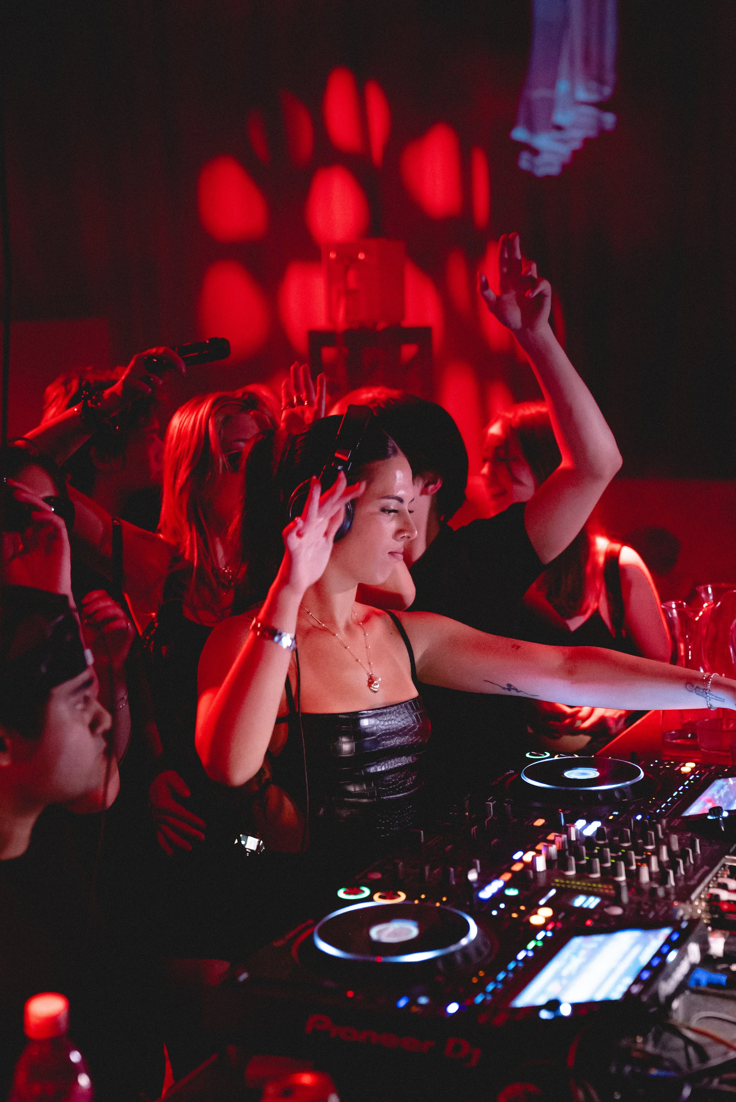 A female DJ with headphones, raising her hand in front of a DJ mixer with colorful lights. Several people are dancing around her in a dark, red-lit environment with a shadowy hand projection on the wall.