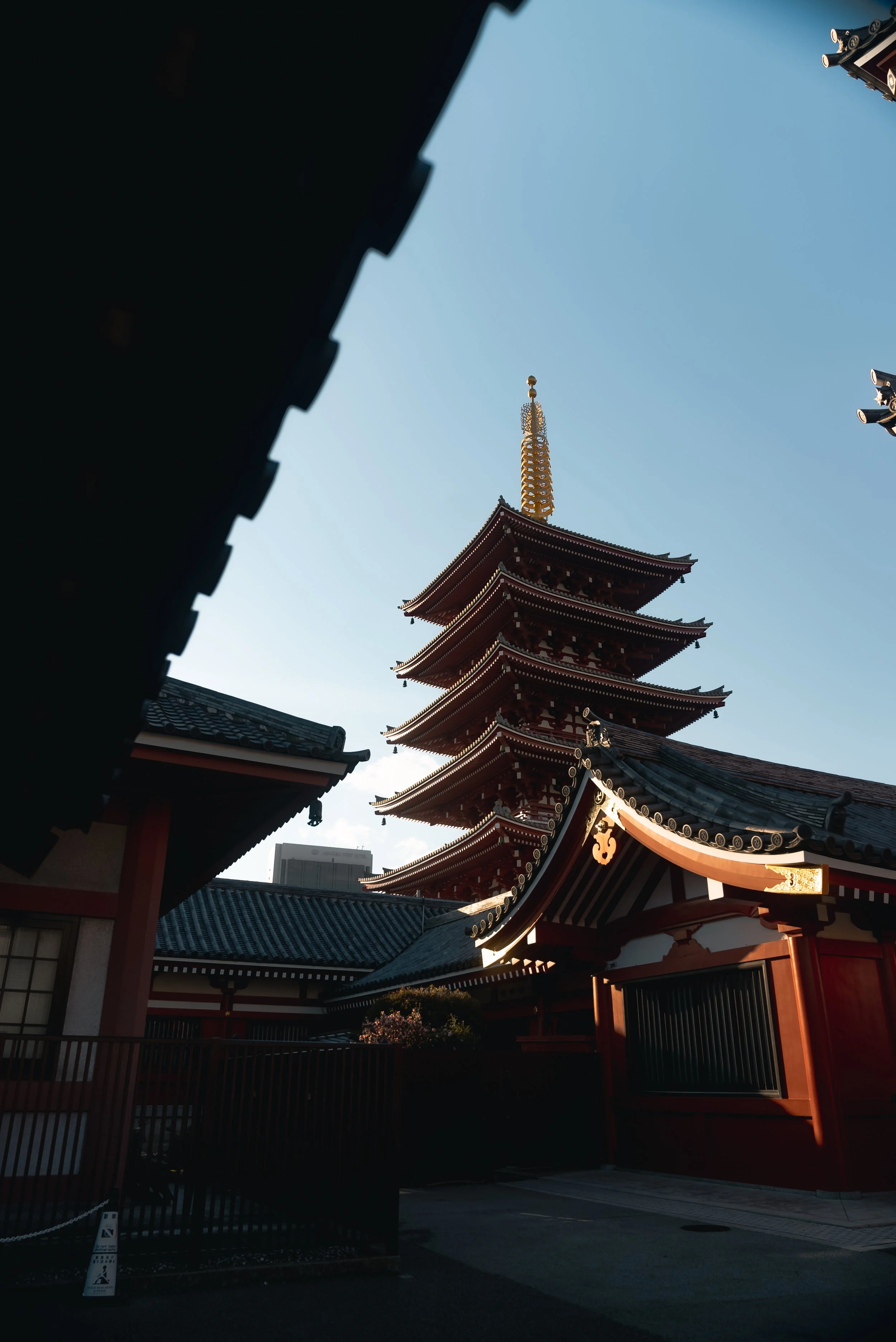 A traditional Japanese pagoda with a multi-tiered roof, ornate eaves, and a golden spire on top, surrounded by other temple buildings, under a clear blue sky.