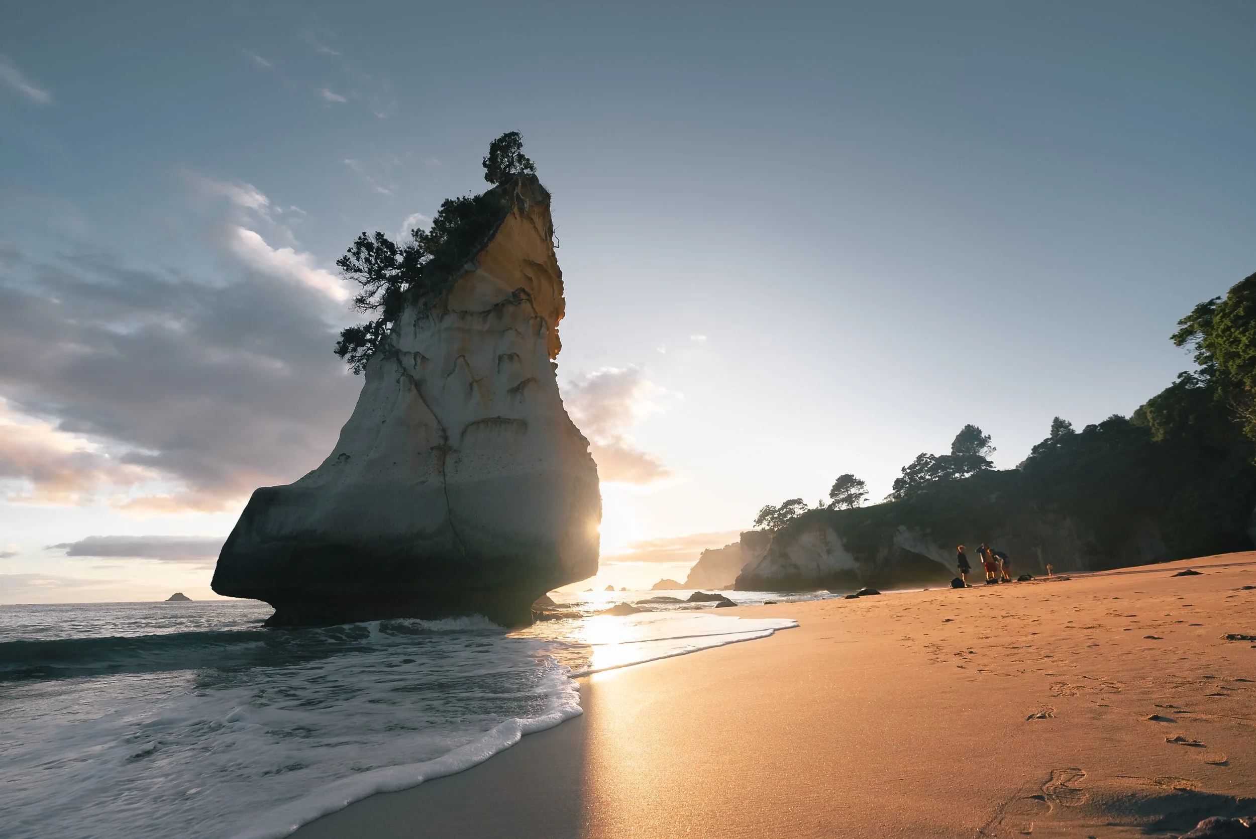 Sunset over a rocky beach with a large rock formation topped with trees, two people walking along the shoreline.