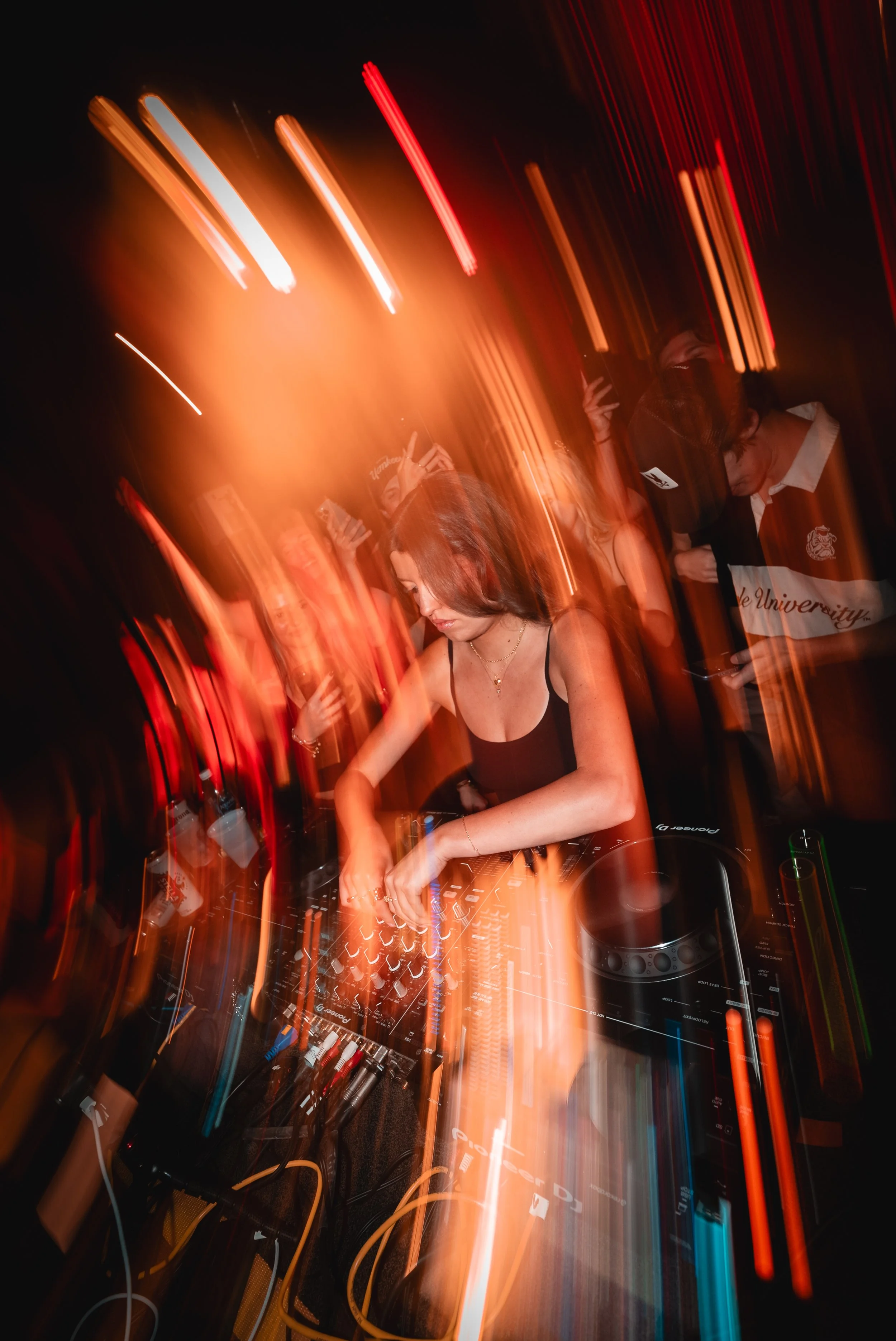 A woman DJ is performing with a Pioneer DJ equipment, surrounded by colorful light streaks creating a dynamic, energetic atmosphere at a nightclub or party.
