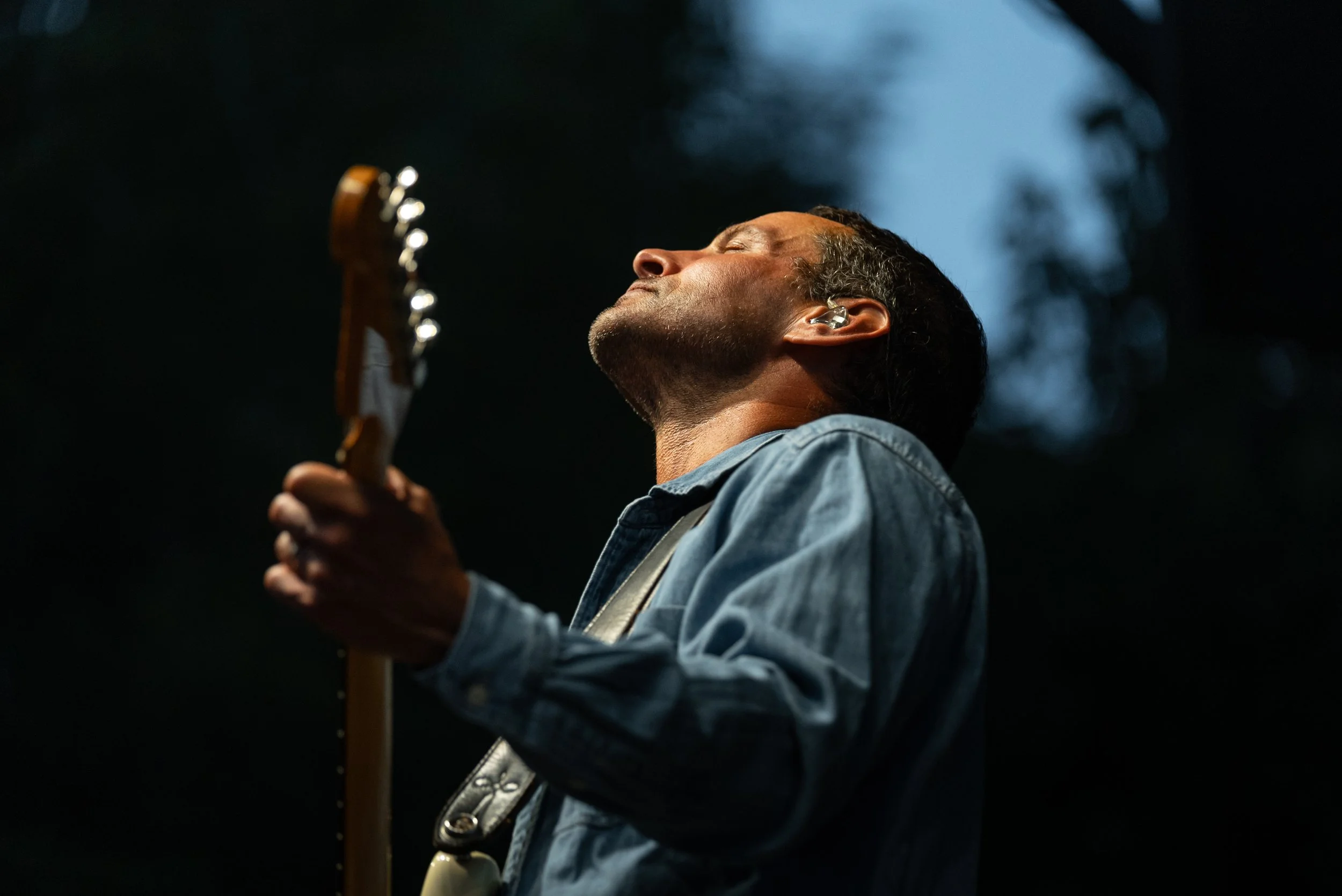 A man with closed eyes playing an electric guitar outdoors at dusk, wearing a denim jacket and an in-ear monitor.
