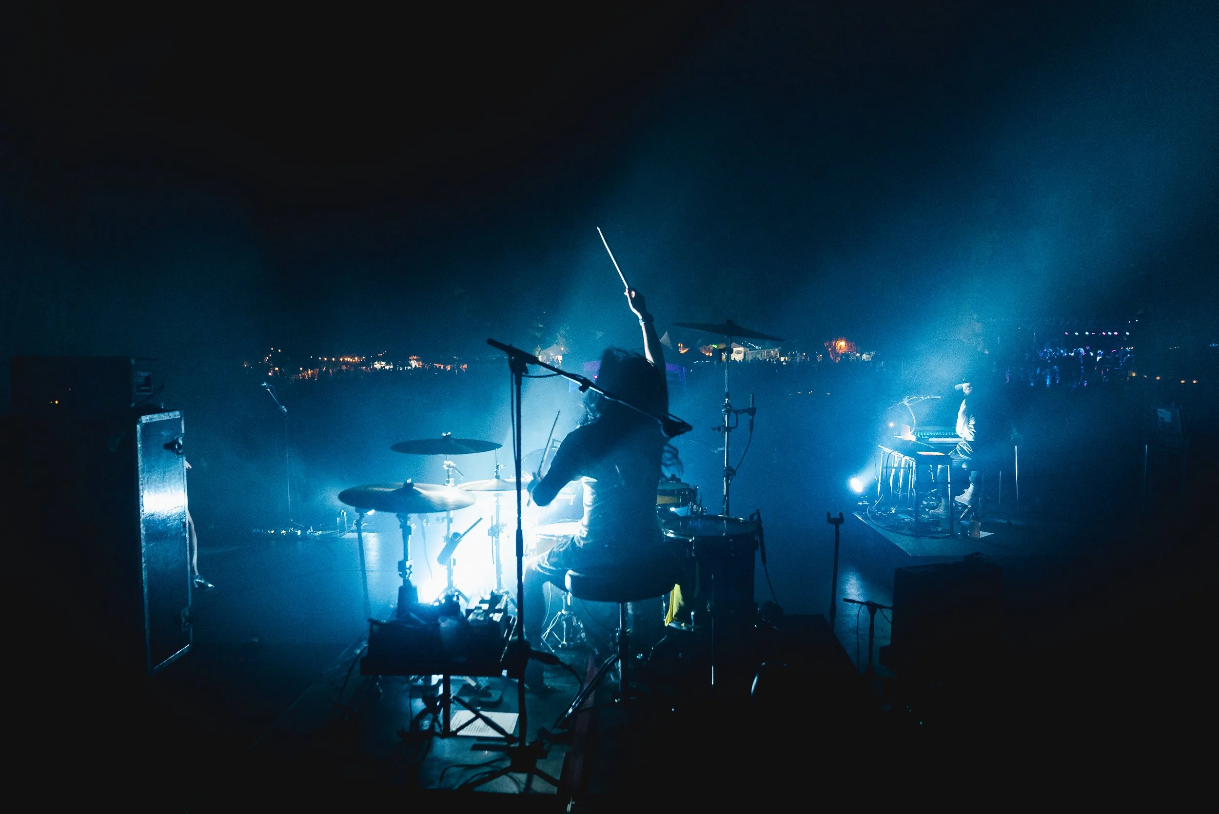 Musicians performing on stage with blue spotlighting at a nighttime outdoor concert, with some audience visible in the background.
