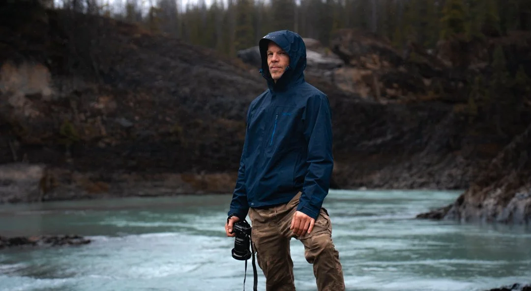 A man in a blue jacket and brown pants standing near a river in a wooded area, holding a camera.