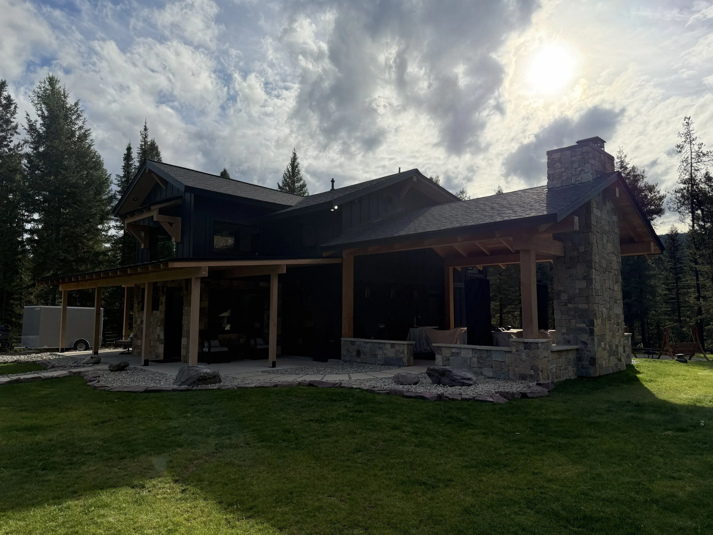 A modern house with a stone chimney, dark exterior walls, and a sloped roof, situated in a wooded area with tall pine trees and a partly cloudy sky.