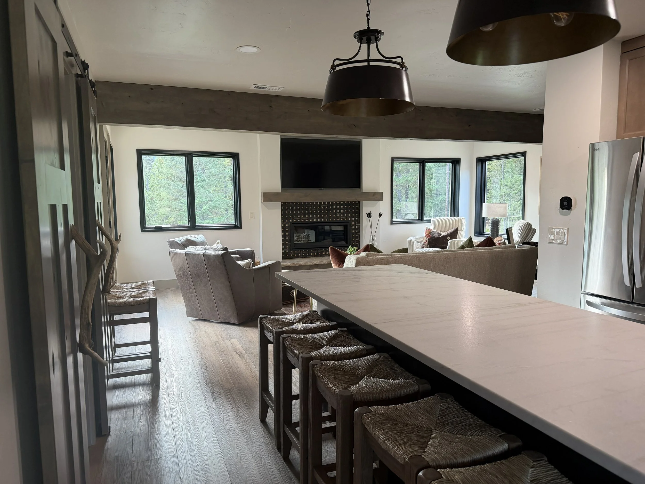 Open-concept living room with large windows, a fireplace, and modern furniture, viewed from the kitchen area with a white countertop and bar stools.