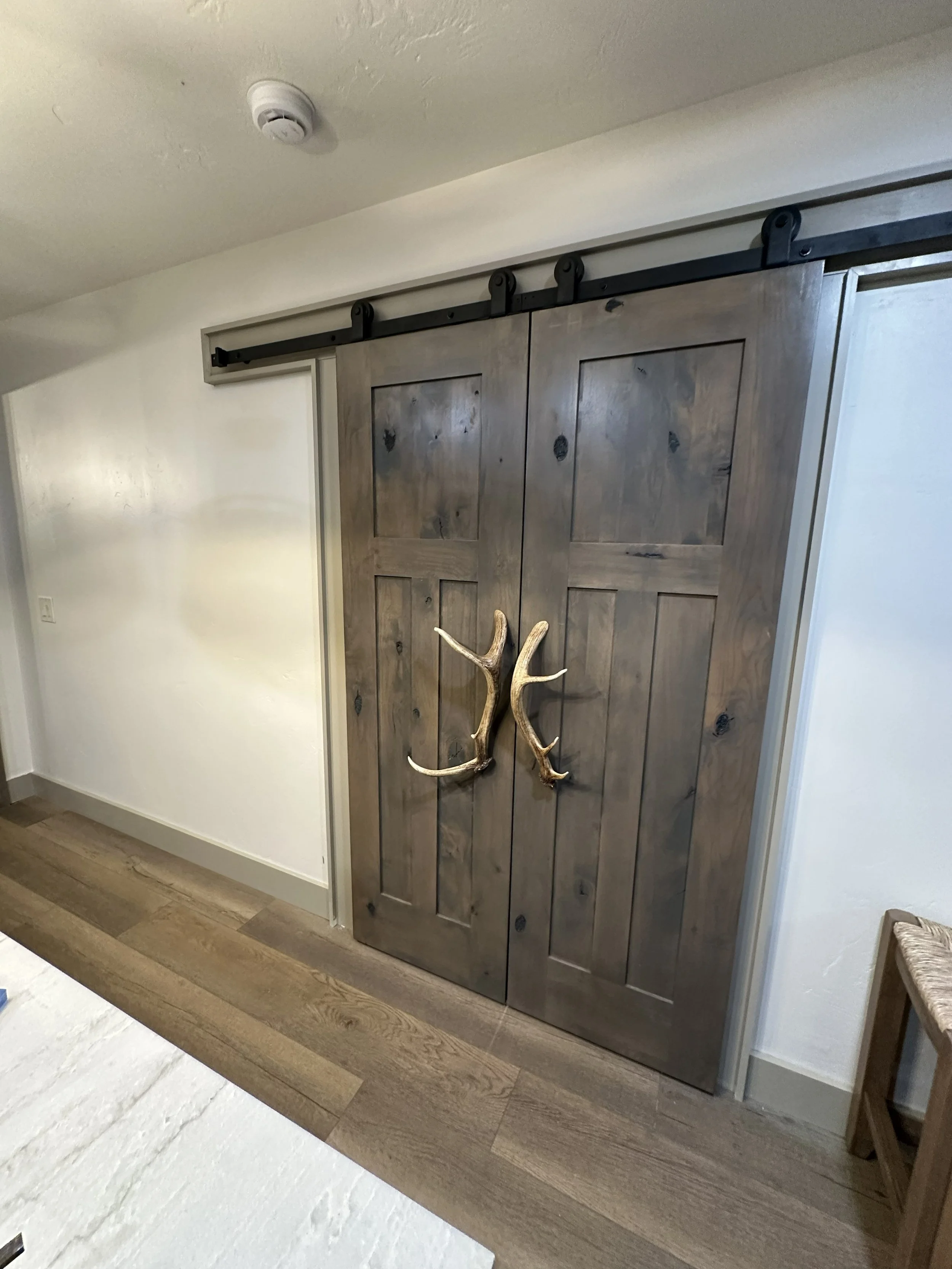 Rustic wooden sliding barn door with black hardware and antler decoration, installed over a white wall in a home interior.