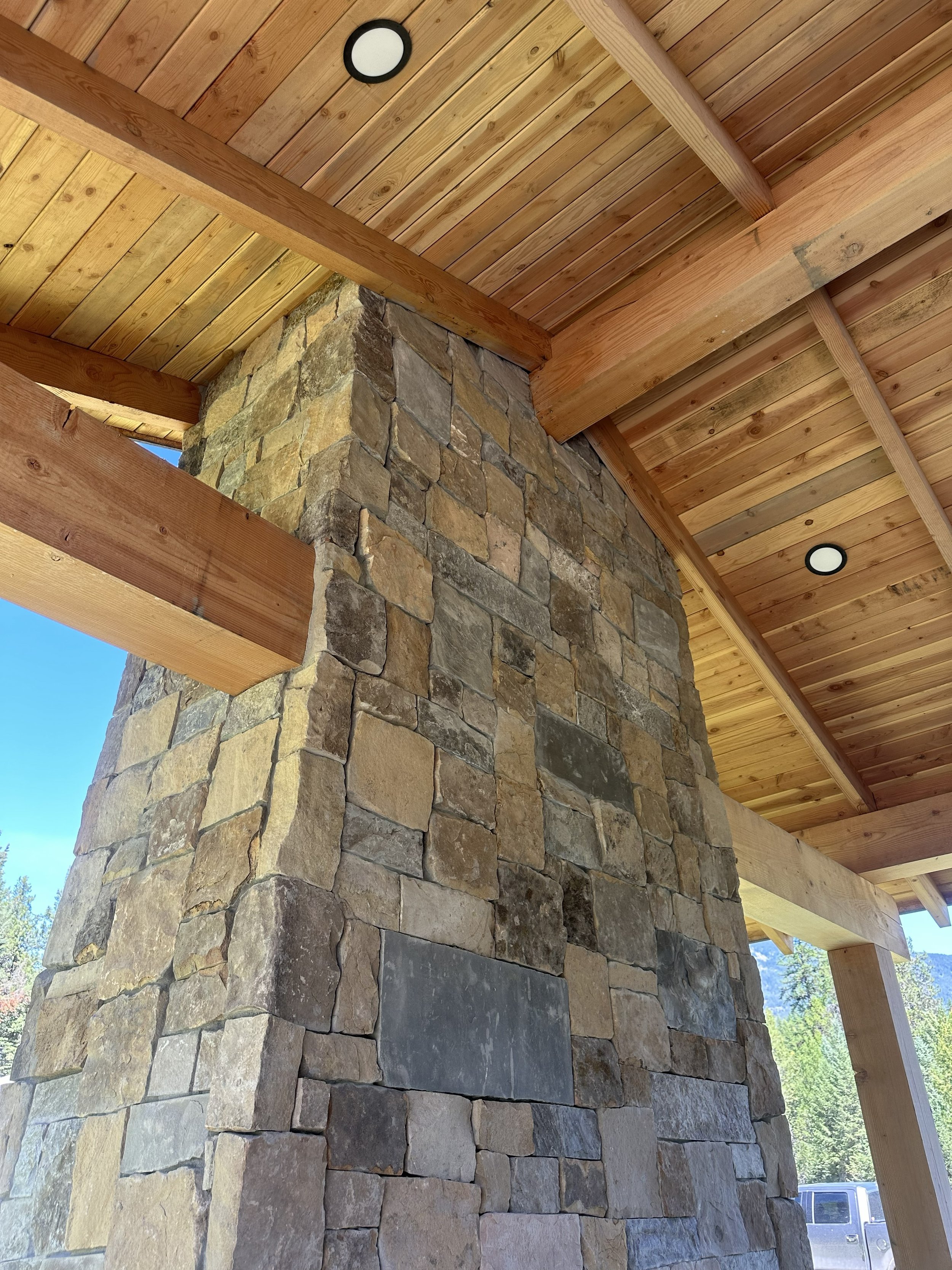 Ceiling with wooden planks and a stone pillar supporting it, two rounds of recessed lighting, and a view of a clear blue sky with trees and a truck outside.