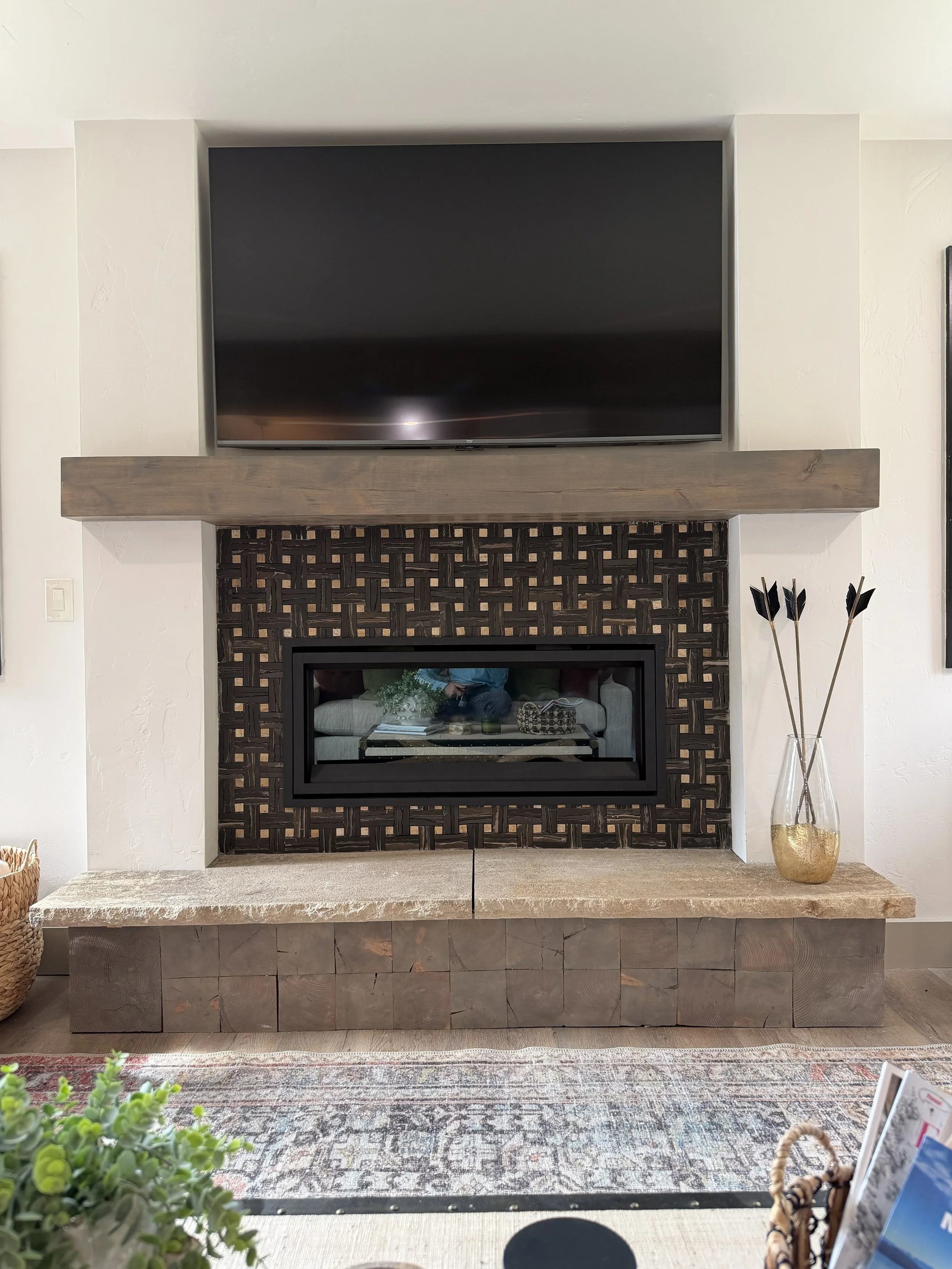 Living room fireplace with a large flat screen TV mounted above a mantel, surrounded by a decorative blackwood lattice, with a vase of black arrows on the side and a woven basket on the floor.