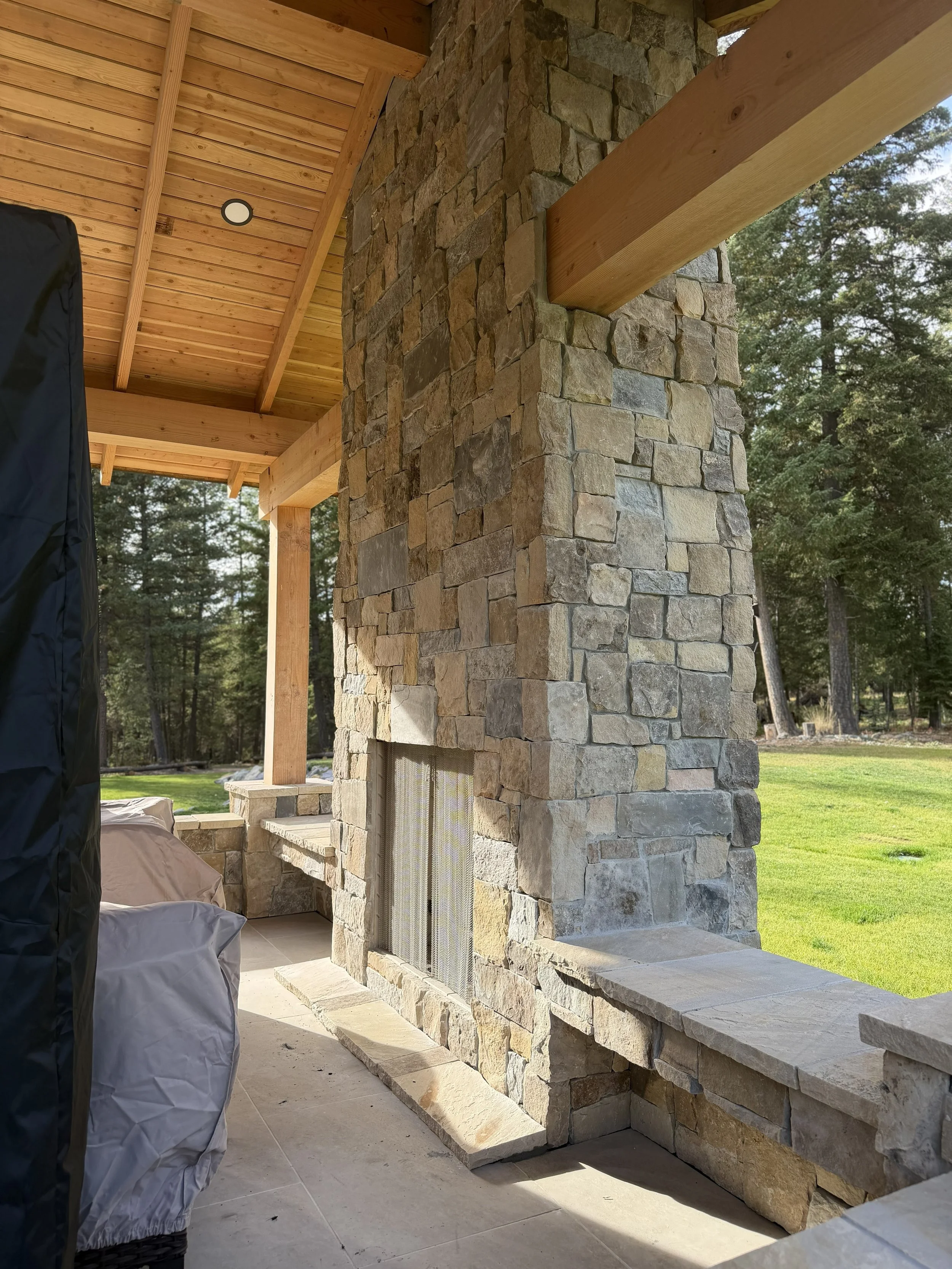 Close-up view of a stone fireplace with a wooden mantel on a patio, with green trees and grass in the background.