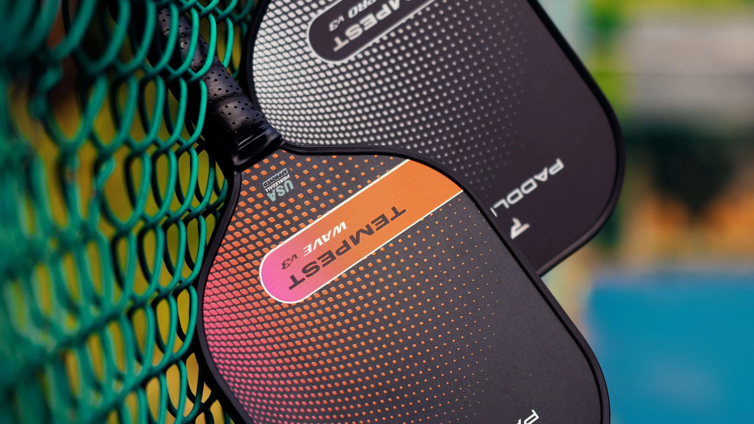PaddleTek Tempest Wave v3 and Reign Pro v3 pickleball paddles hanging on chain-link fence at outdoor court