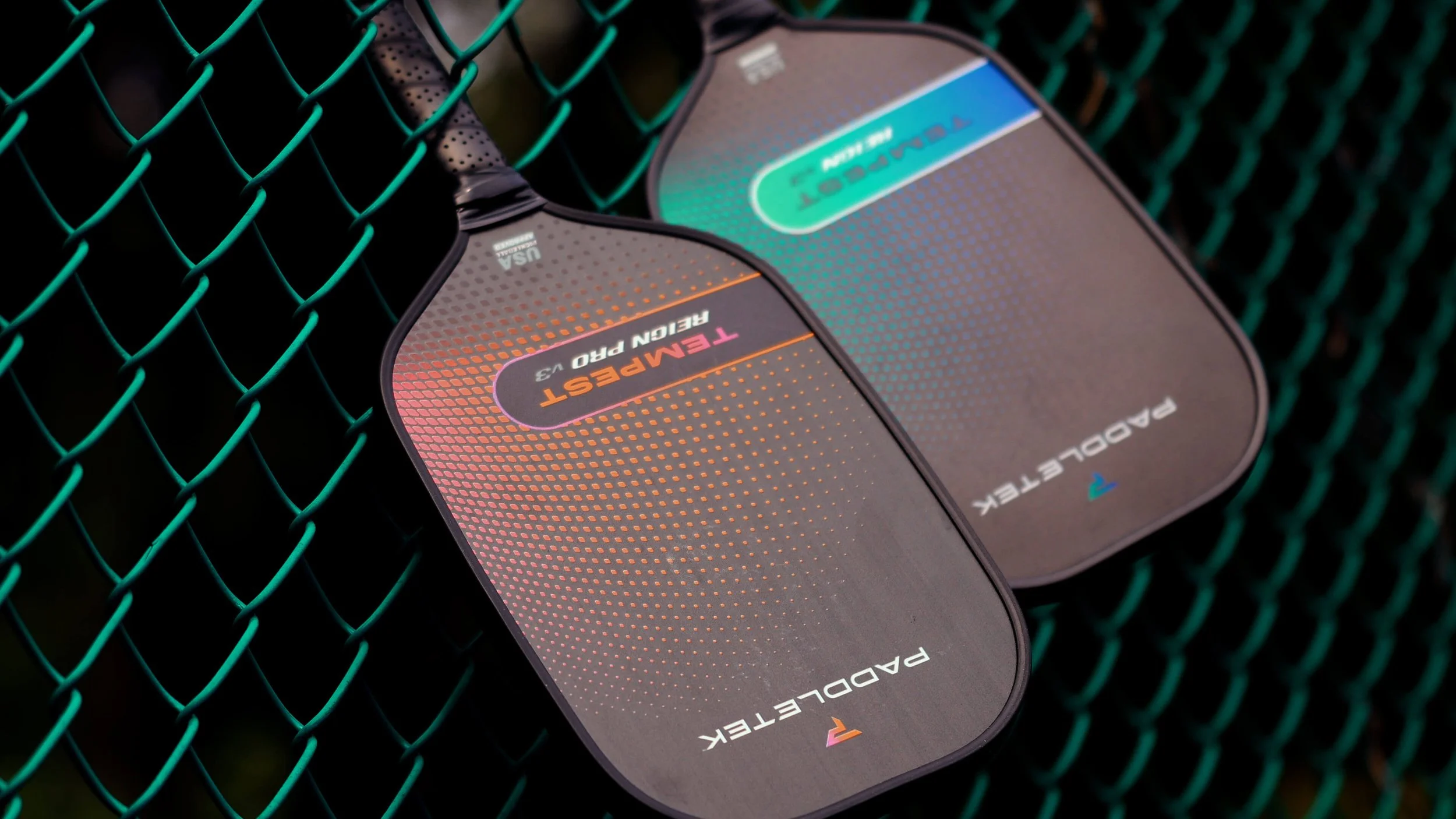 PaddleTek Tempest Reign Pro v3 and Tempest Reign v3 pickleball paddles leaning on chain-link fence