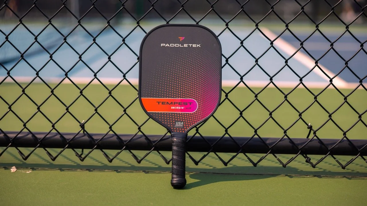 PaddleTek Tempest Wave v3 pickleball paddle leaning against chain-link fence on outdoor court