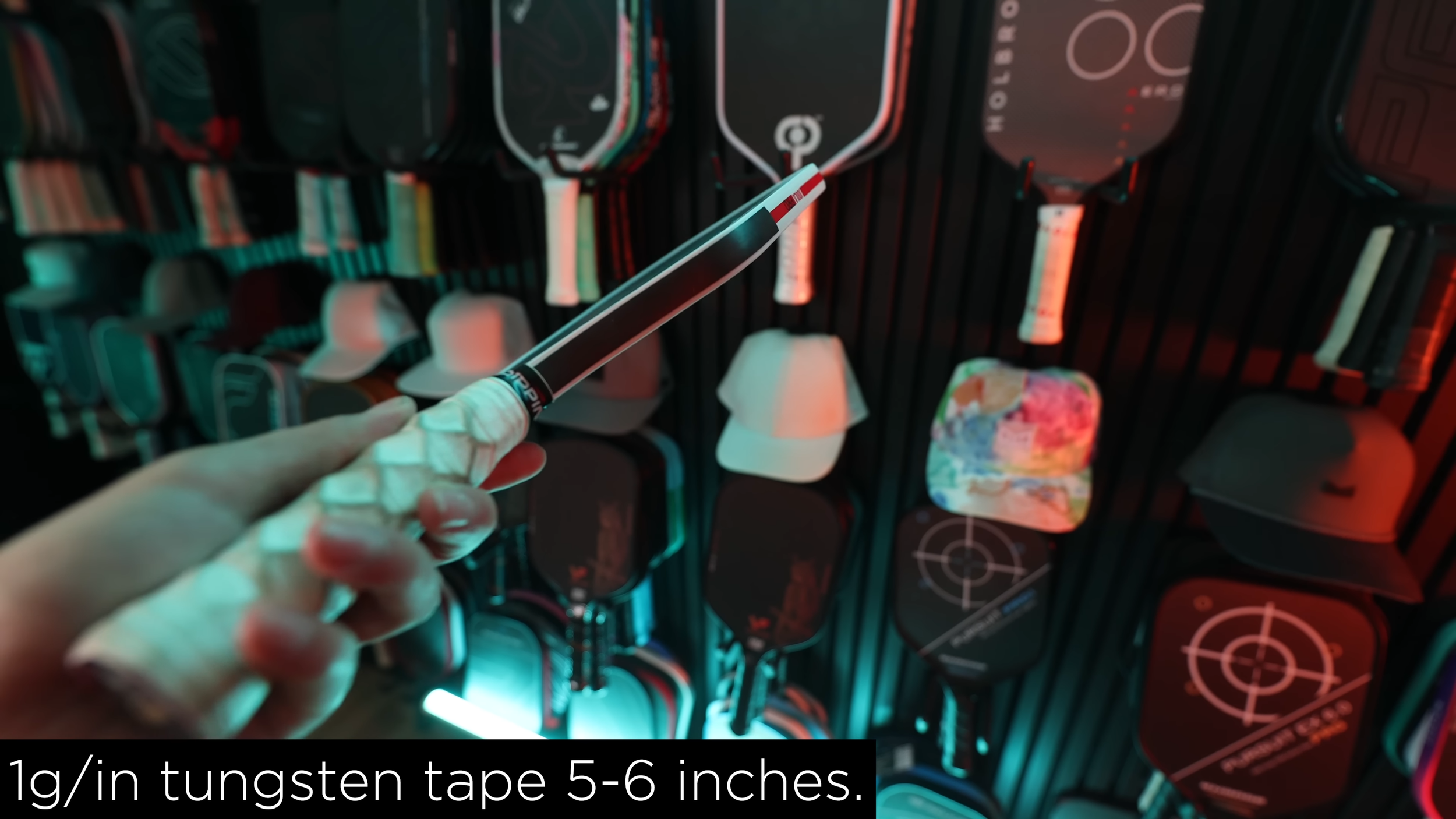 Tungsten tape being applied to a pickleball paddle handle at 1g per inch for 5-6 inches in a studio with paddle wall