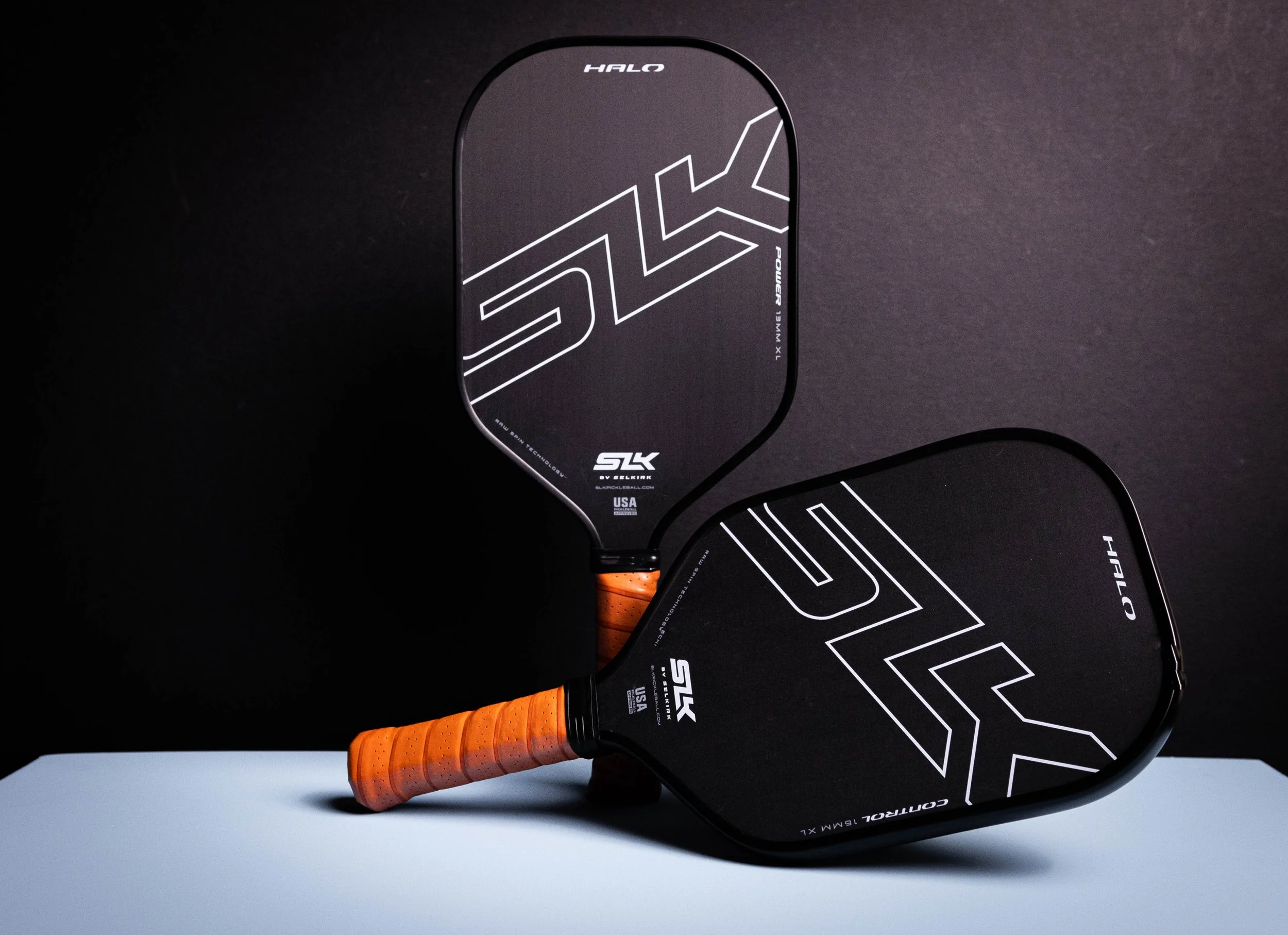 Two SLK Halo Control XL pickleball paddles with black faces and orange grips displayed against a dark background