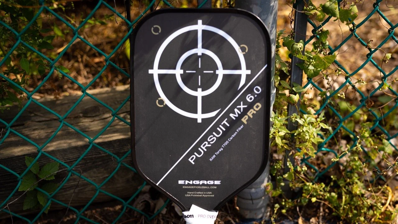 Engage Pursuit MX 6.0 Pro pickleball paddle leaning against a chain-link fence on an outdoor court