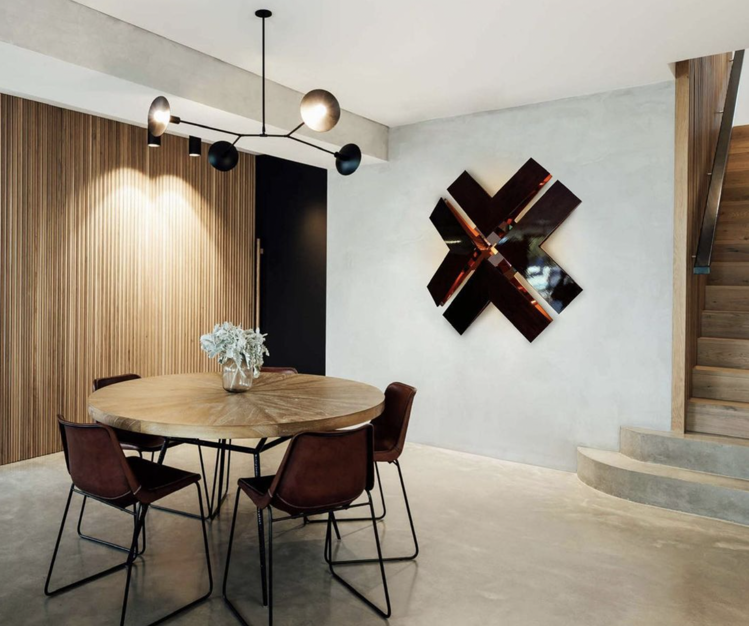 Modern dining area with round wooden table, brown chairs, contemporary chandelier, wall art, wood-paneled wall, and staircase.