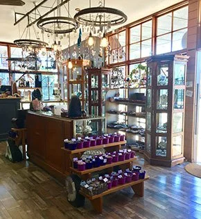 Display of packaged food items on wooden shelves inside a well-lit shop with large windows and decorative hanging light fixtures.