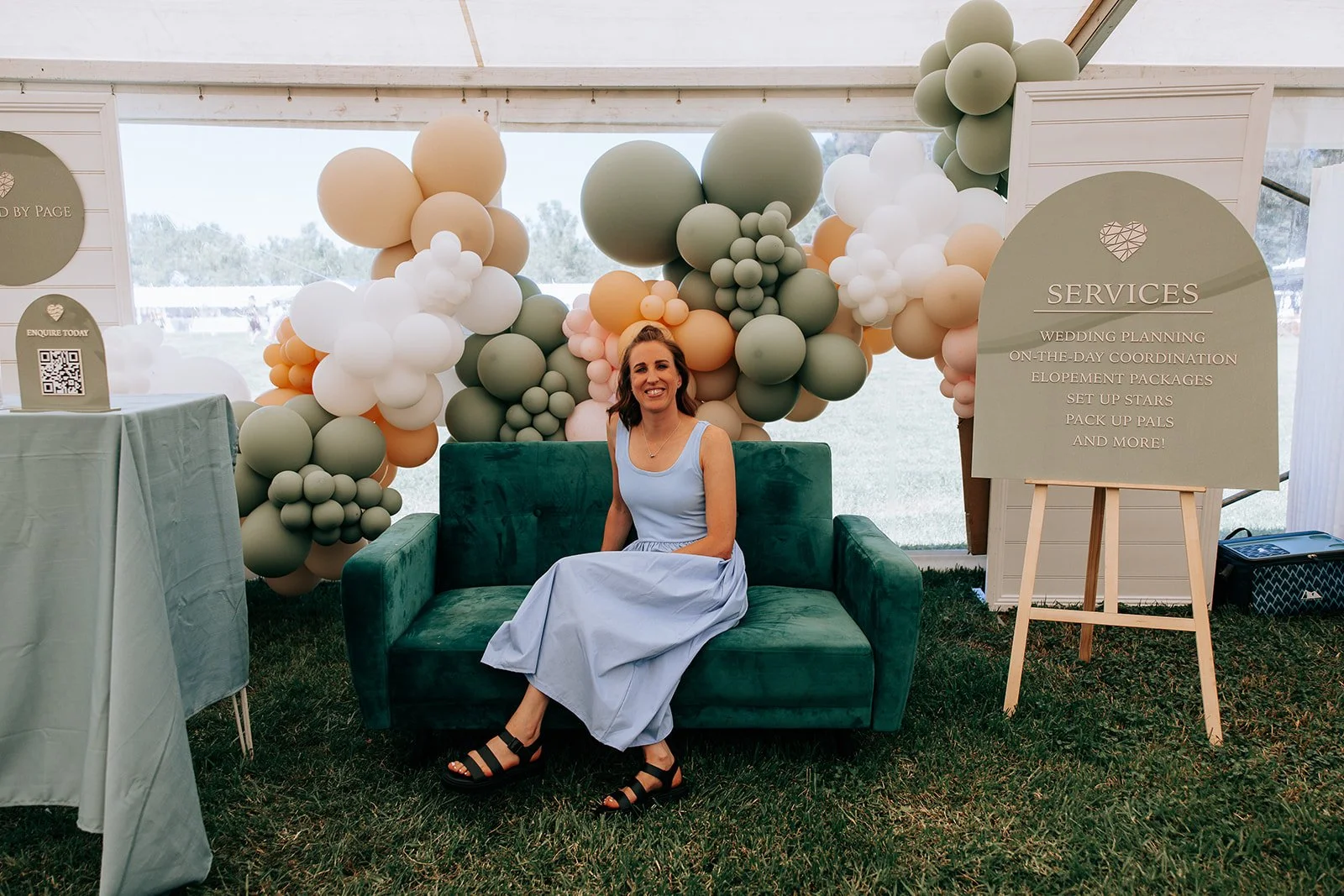 Woman sitting on a green couch inside a tent, surrounded by pastel balloon decorations, with a sign listing wedding services like planning and coordination.