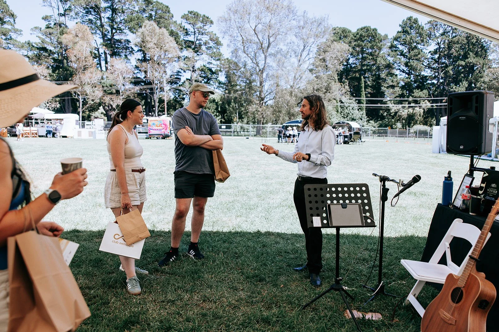 Group of people standing outdoors at a casual event, interacting near musical equipment and holding shopping bags.