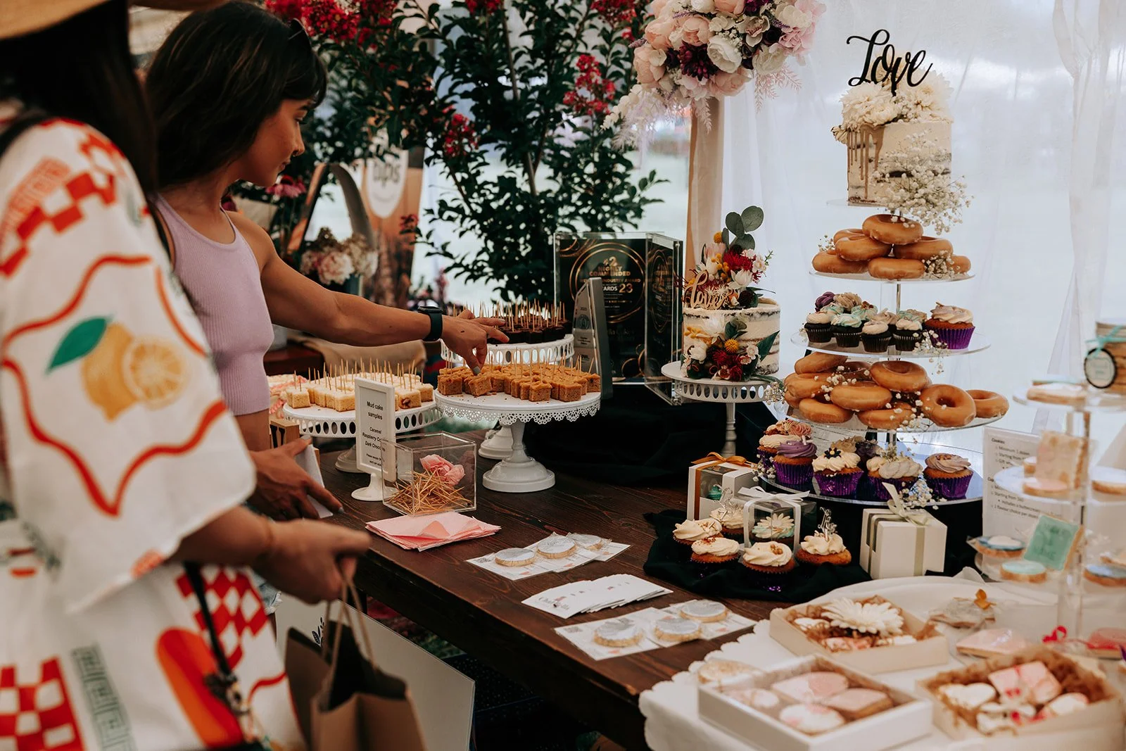 People browsing a dessert table with cakes, donuts, cupcakes, and cookies, decorated with floral arrangements and 'Love' cake topper.