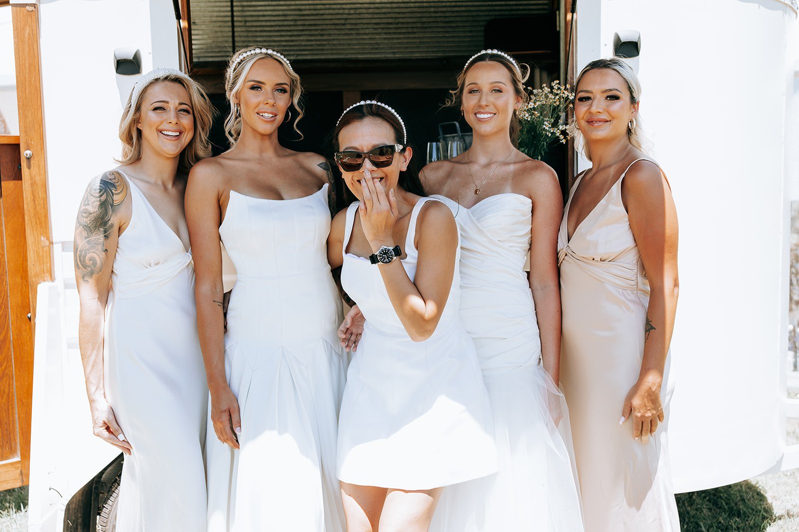 Five women in white dresses, smiling and posing in front of a trailer, with one wearing sunglasses and jewelry.