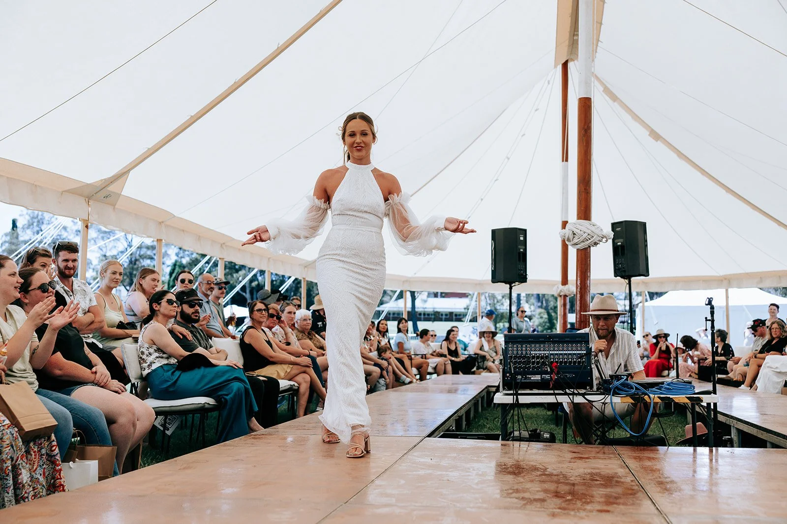 A person in a white dress walking on a runway at a fashion show under a large tent, surrounded by an audience seated on either side, some clapping.