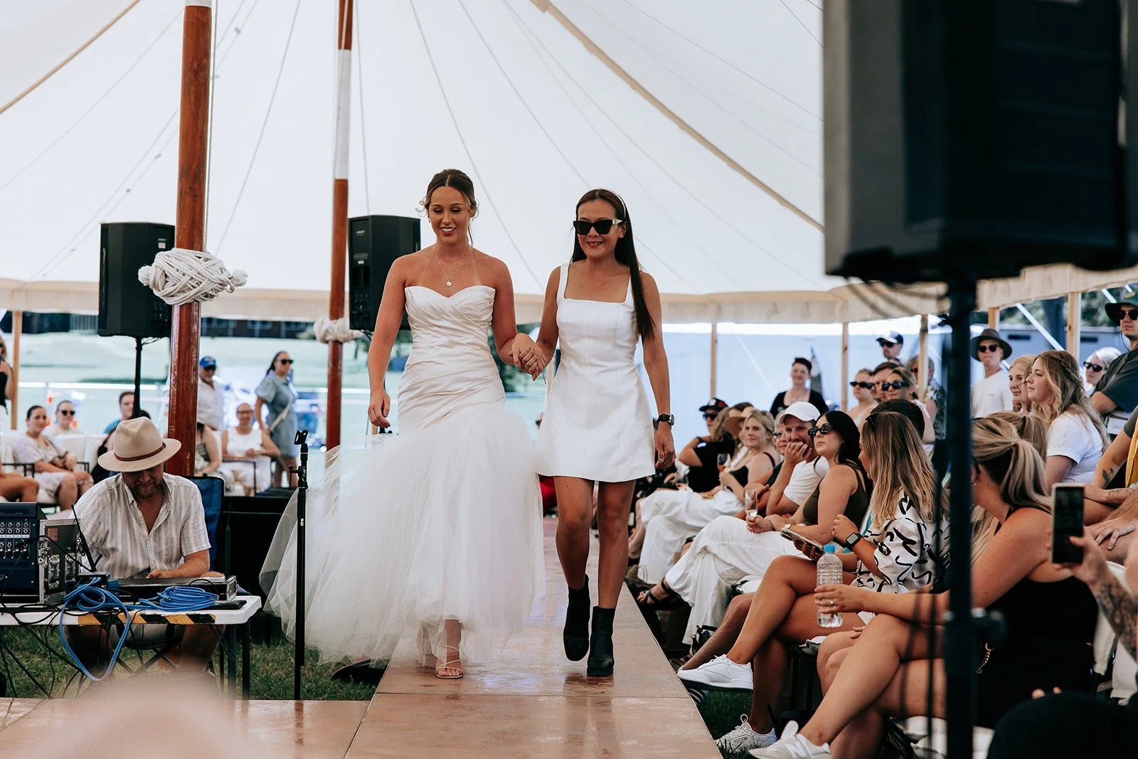 Women walking on runway at fashion show tent with audience seated around.