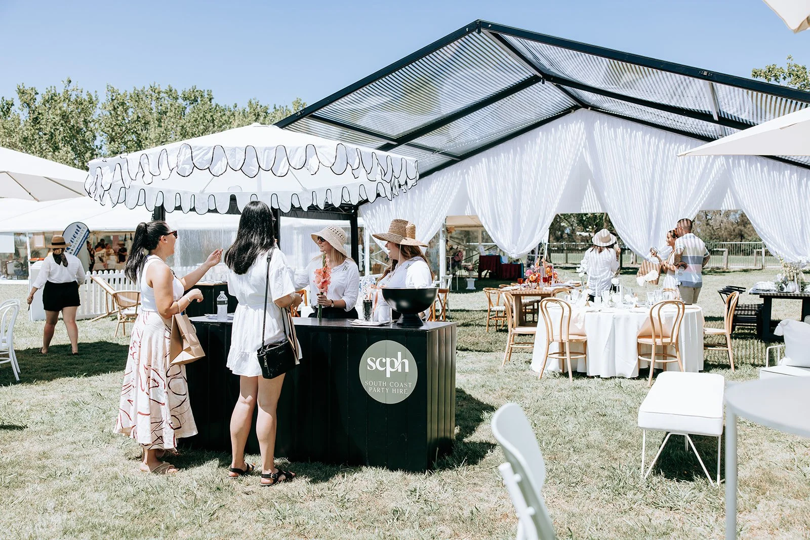 Outdoor event with people under a white tent with tables and umbrellas, interacting near a black bar labeled "South Coast Party Hire."