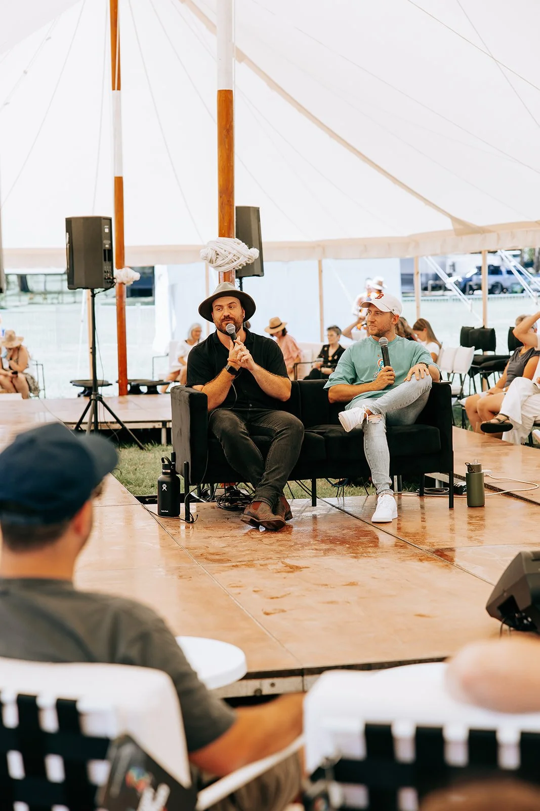 Two men sitting on a stage in a tent, holding microphones, surrounded by an audience. One man wears a wide-brimmed hat. Chairs and sound equipment are visible.