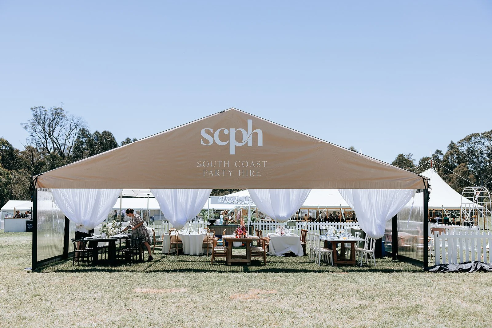 Outdoor event tent labeled "South Coast Party Hire" with tables and chairs set up underneath, surrounded by grassy area and additional tents in the background.