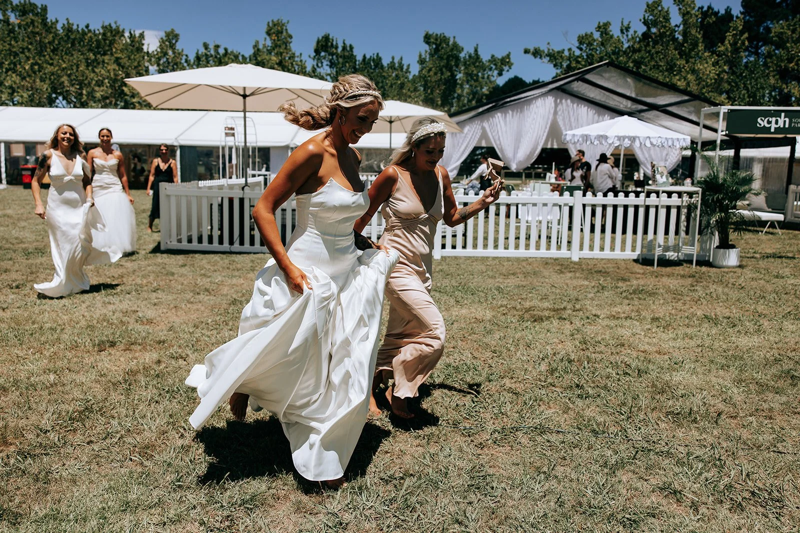 Women in dresses running on grass at an outdoor wedding reception with tents and umbrellas in the background.