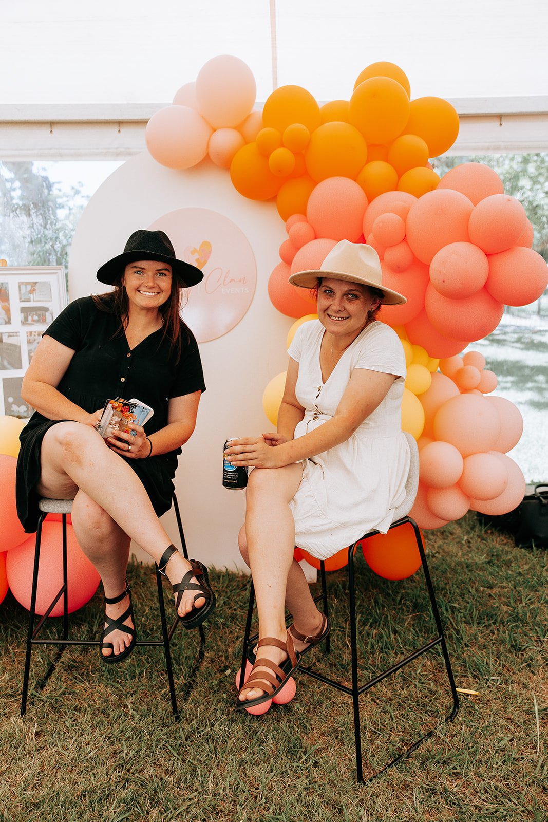 Two women sitting on stools in front of a balloon display, each holding canned drinks, wearing hats and dresses, in an outdoor setting.