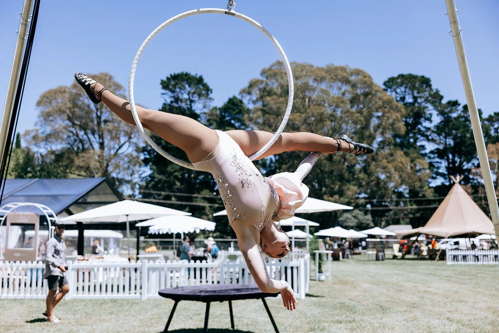 Aerial performer in a pastel costume executing a pose on a hoop with outdoor event backdrop.