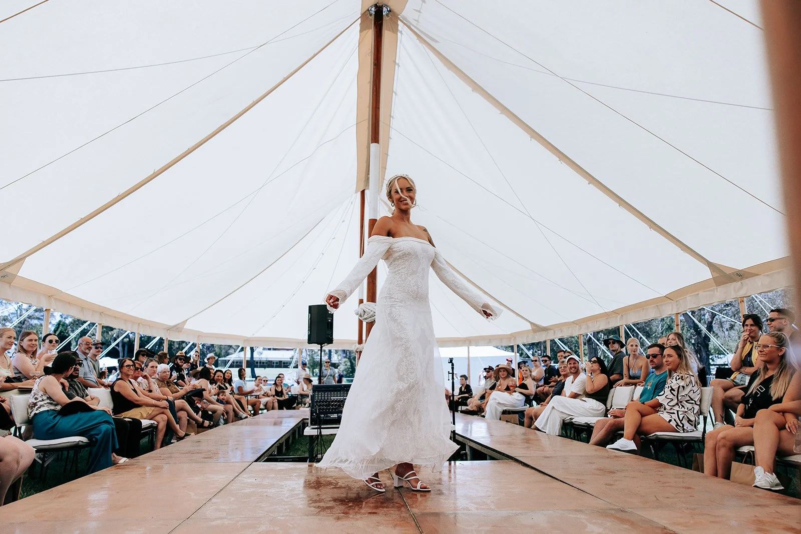 A model wearing a long white off-the-shoulder dress walks on a wooden runway under a large tent, surrounded by seated spectators.