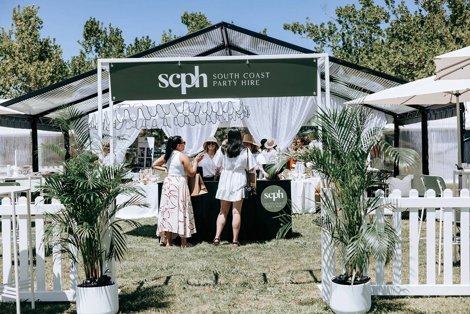 Outdoor event setup with South Coast Party Hire signage, featuring a white picket fence, potted plants, and a group of people beneath a tent with tables and decorations.