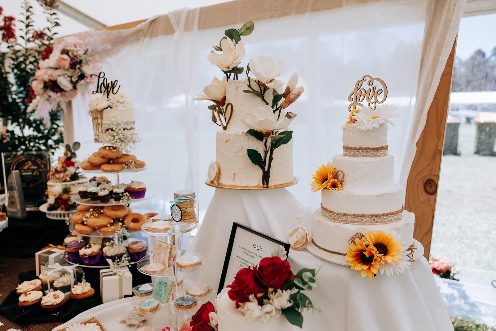 A wedding dessert table featuring two decorated cakes with floral accents and "love" toppers, an assortment of donuts, cupcakes, and cookies on display stands.