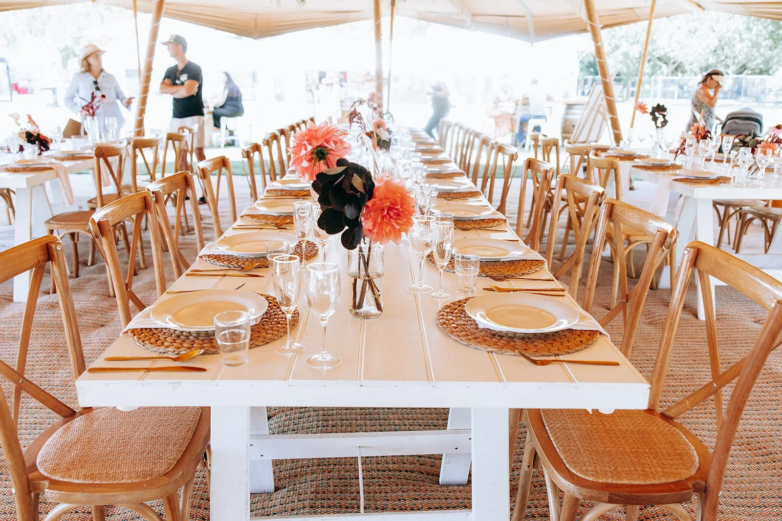 Long dining table set for an event with wooden chairs, woven placemats, white plates, glassware, and floral centerpieces under a canopy.