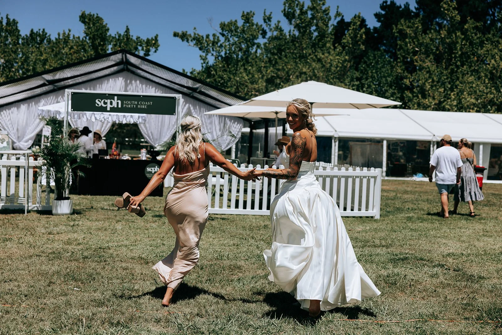A woman in a wedding dress and another woman in a light dress joyfully running on grass towards a party hire tent by scph, with a white picket fence and trees in the background.