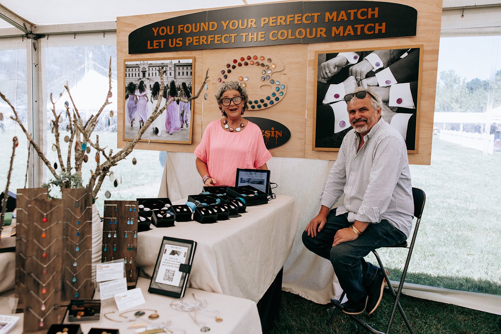 Two people at a jewelry booth with custom color-matching products displayed, featuring necklaces and bracelets on a table. A sign above reads, 'You found your perfect match, let us perfect the color match.'