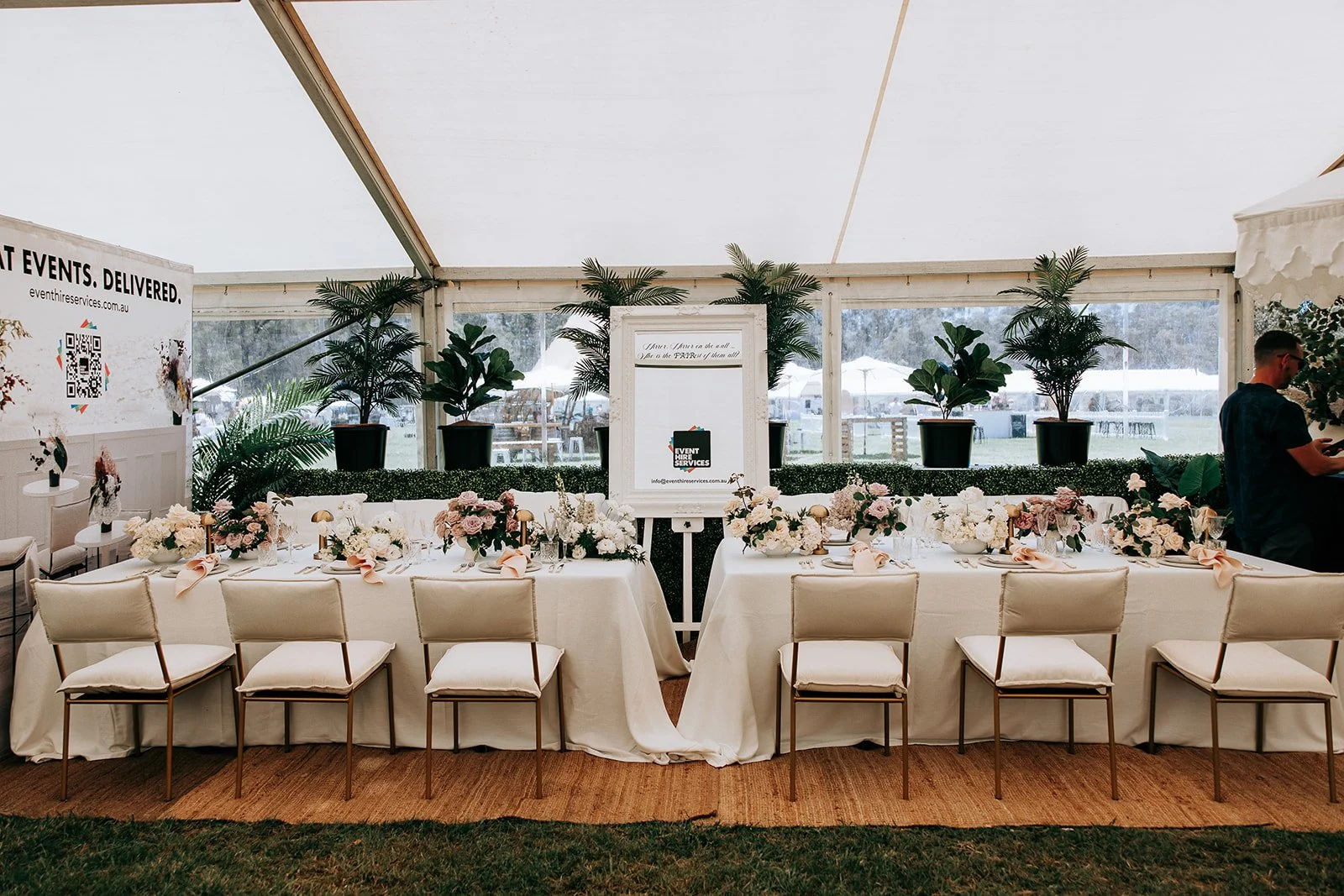 Elegant event setup with a long table covered in a white tablecloth, adorned with floral centerpieces and neatly arranged tableware, surrounded by chairs. Potted plants are displayed along the backdrop, and a framed sign with contact information is p